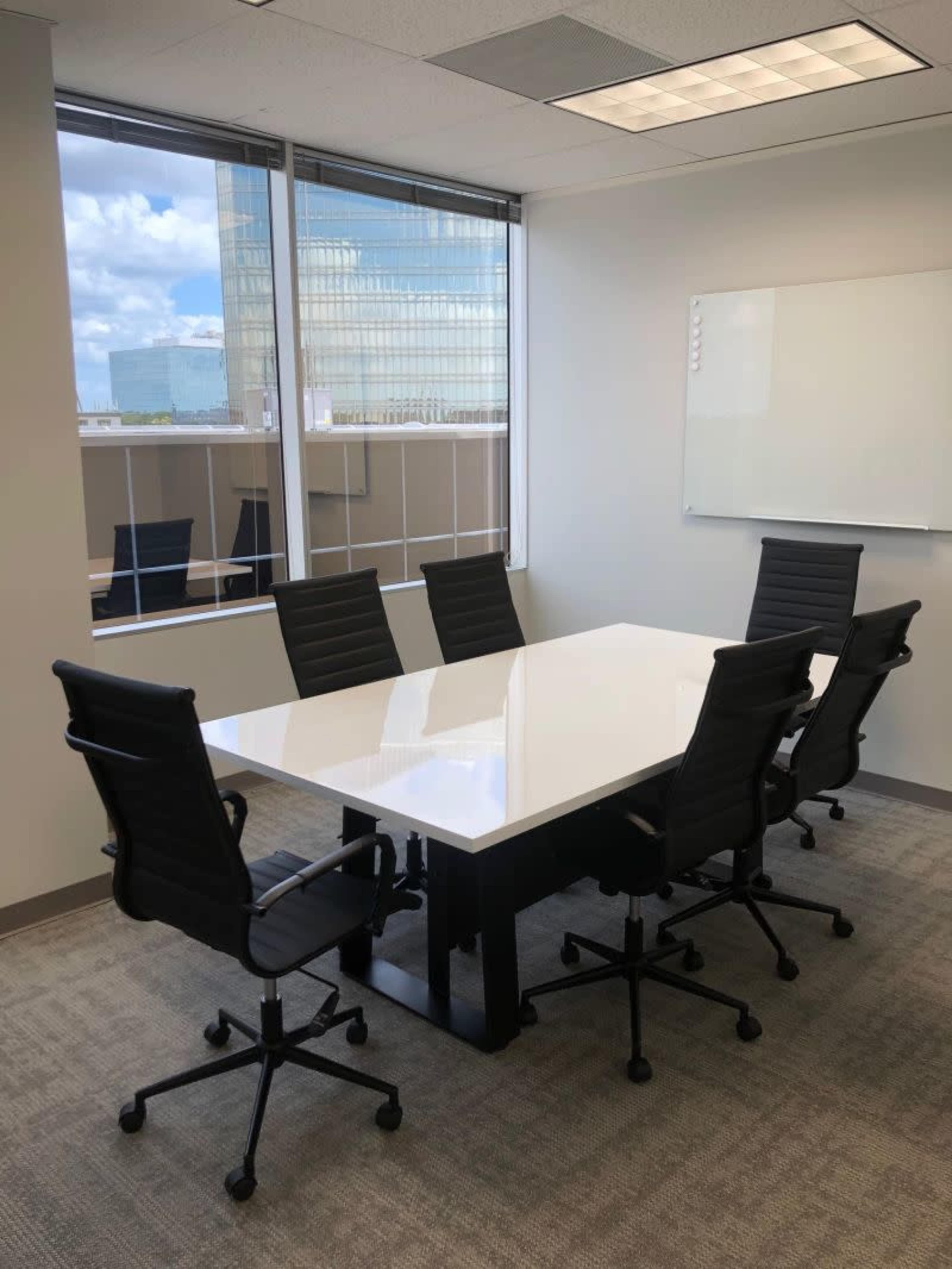 A modern conference room with a large white table and six black chairs surrounded by windows.