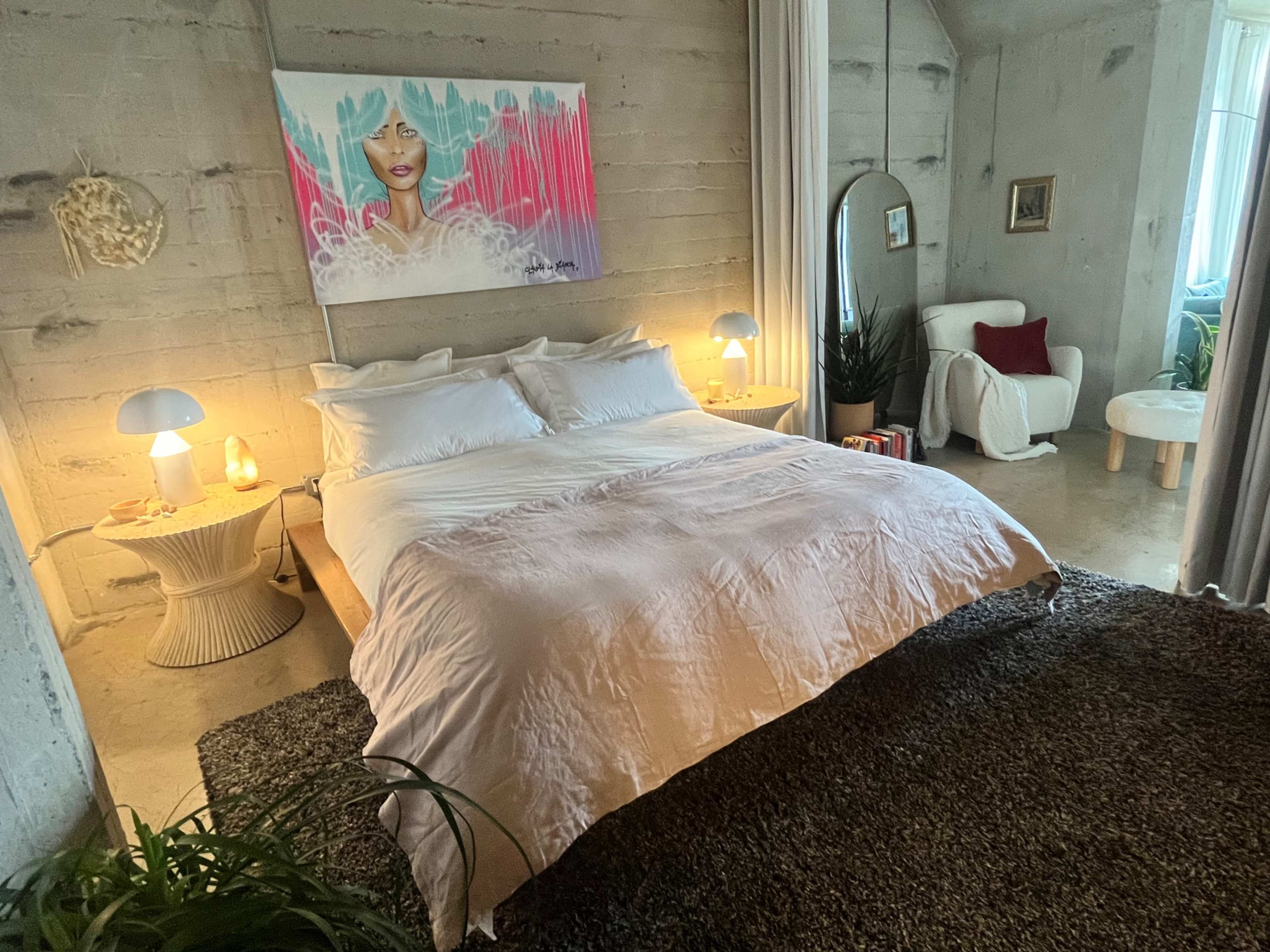 The scene shows a stylish bedroom featuring a large bed with white bedding, a colorful artwork on the wall, and warm lighting from table lamps.