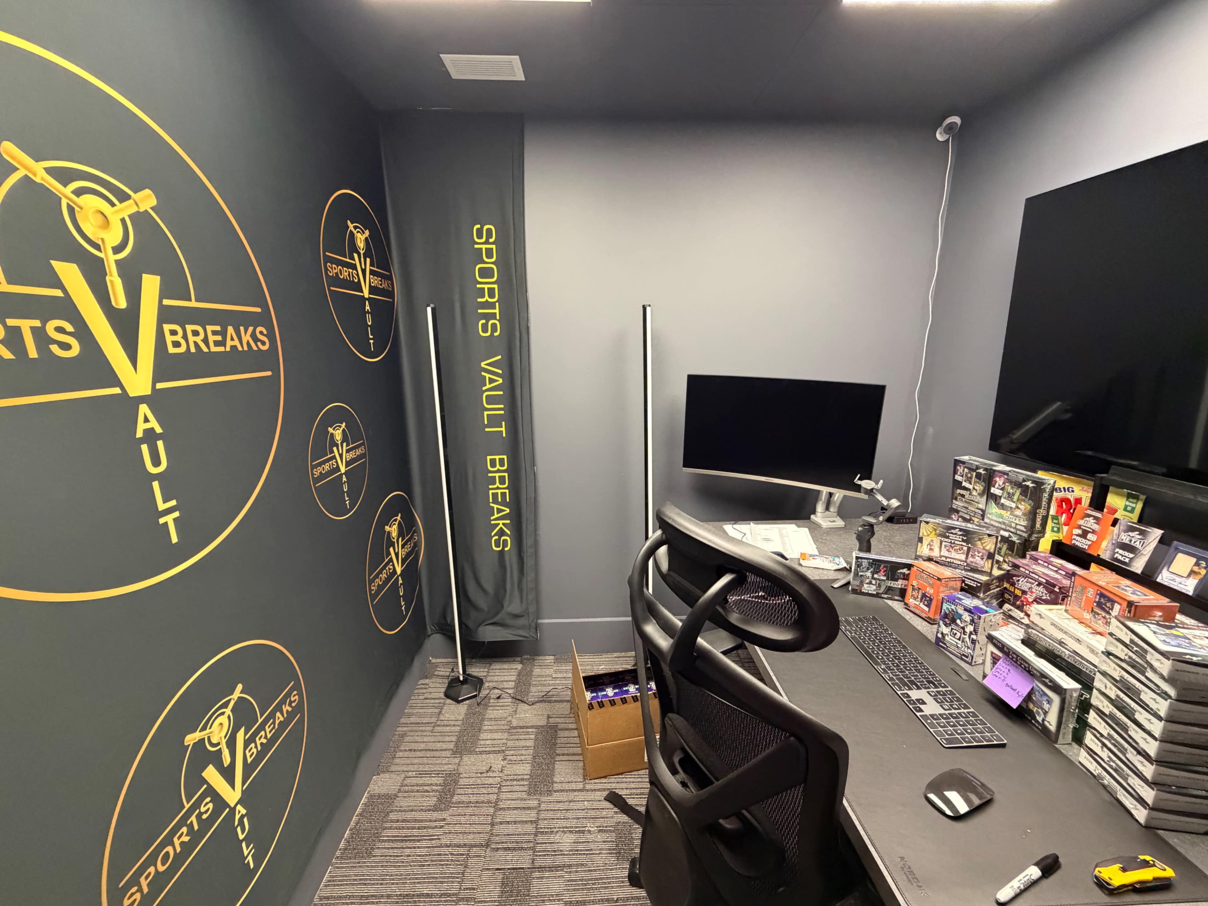 The image shows a small office or workspace designed for sports card breaks, featuring a desk with a computer monitor, shelves of packaged sports cards, and branded banners on the walls.