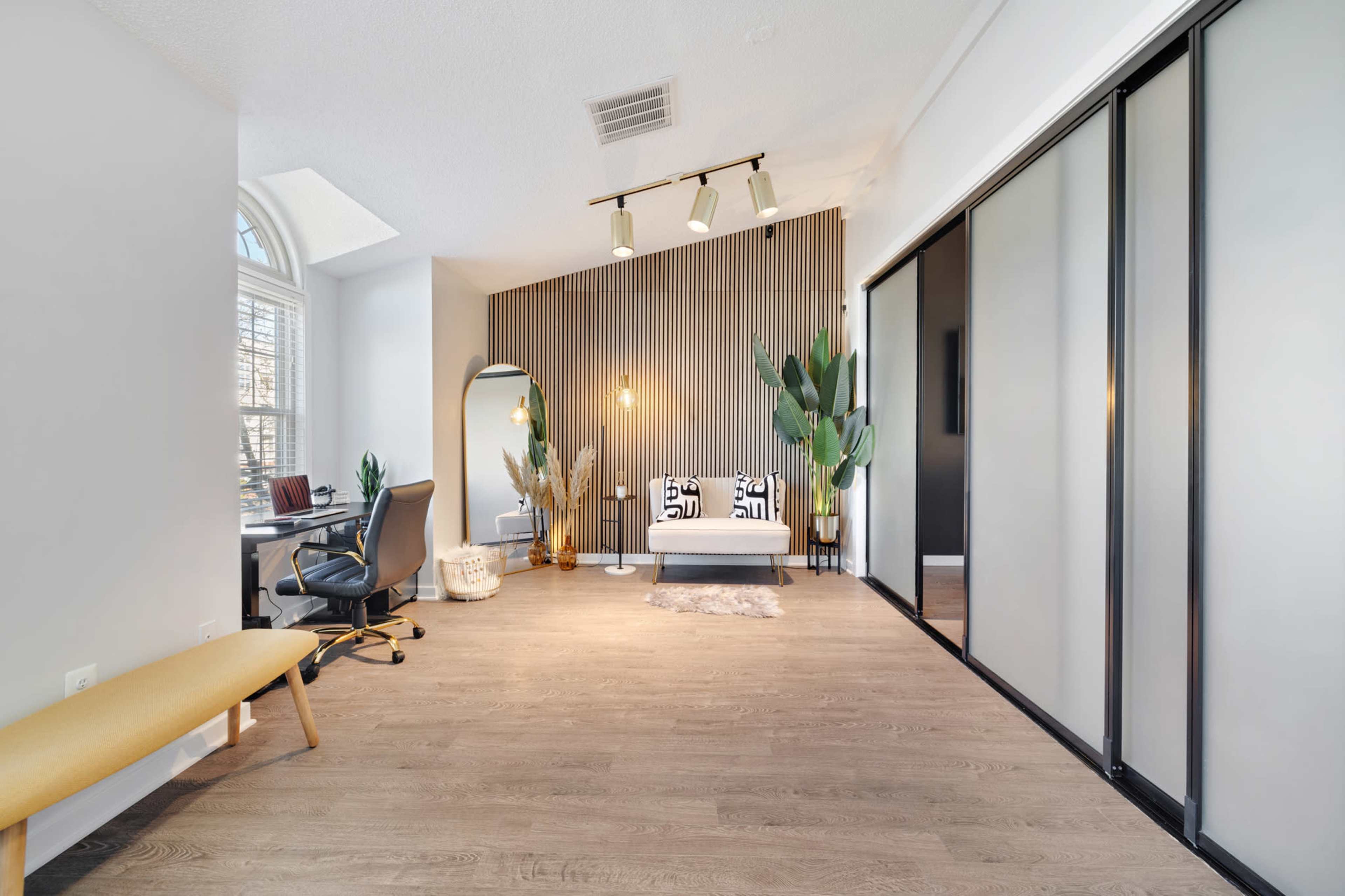 A modern office space features a seating area with a sofa and plants, complemented by a desk with a chair and large windows allowing natural light.