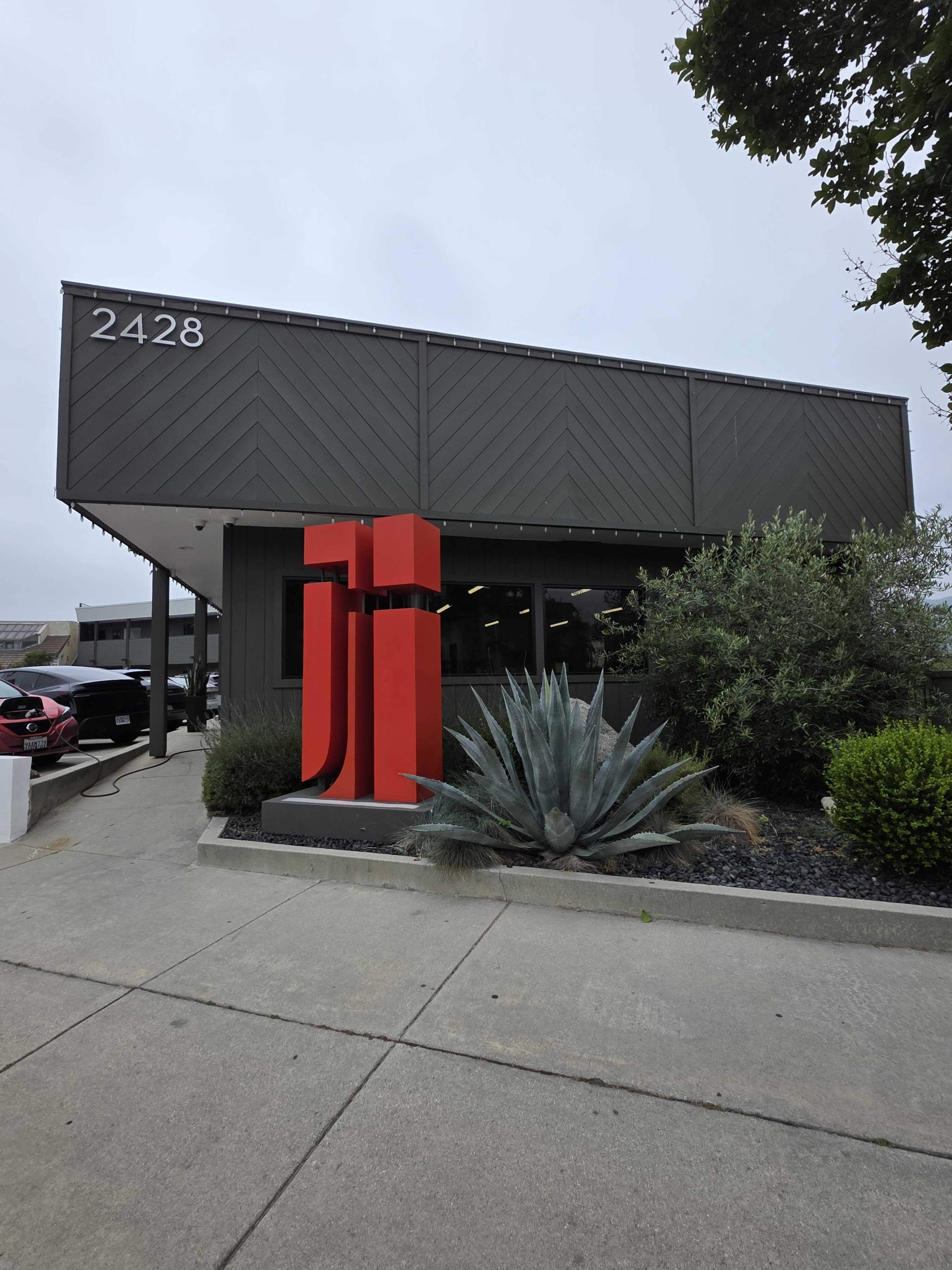 The image shows a modern building with a large red sign displaying "JJ" in front of landscaped greenery and a concrete walkway.