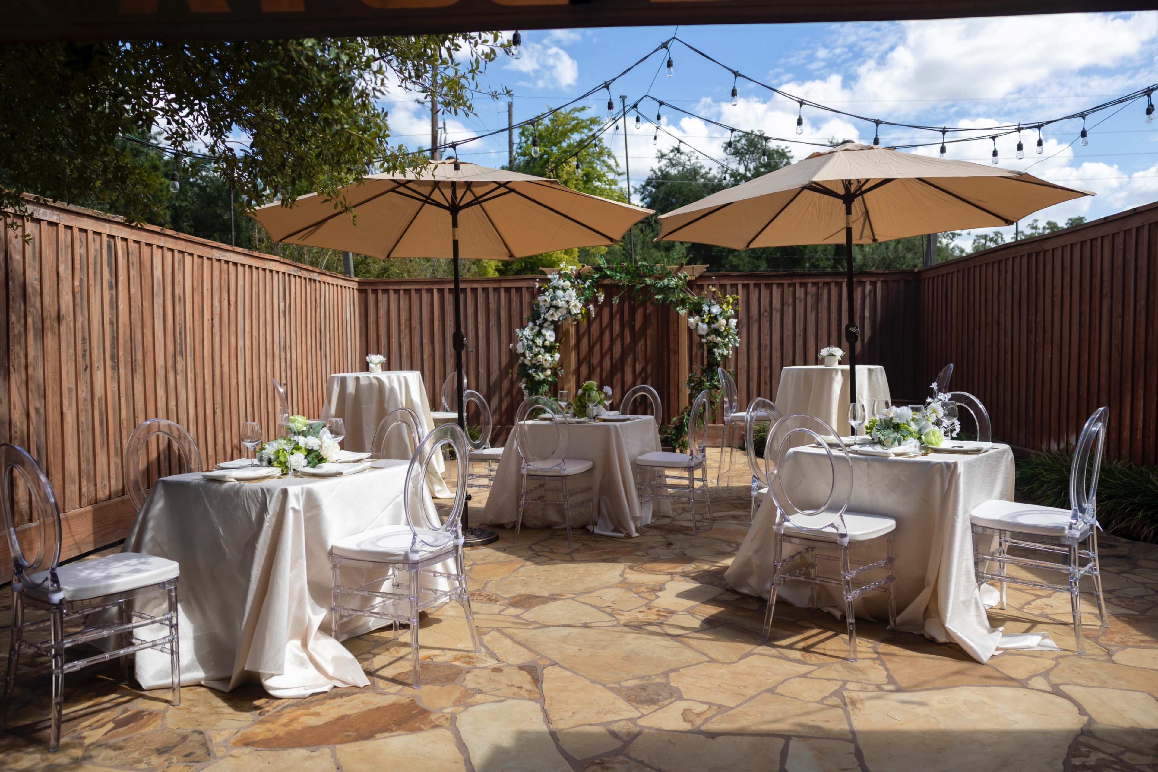 The image shows an outdoor event space with several round tables covered in white tablecloths, clear chairs, and two large umbrellas, surrounded by wooden fencing and decorative greenery.