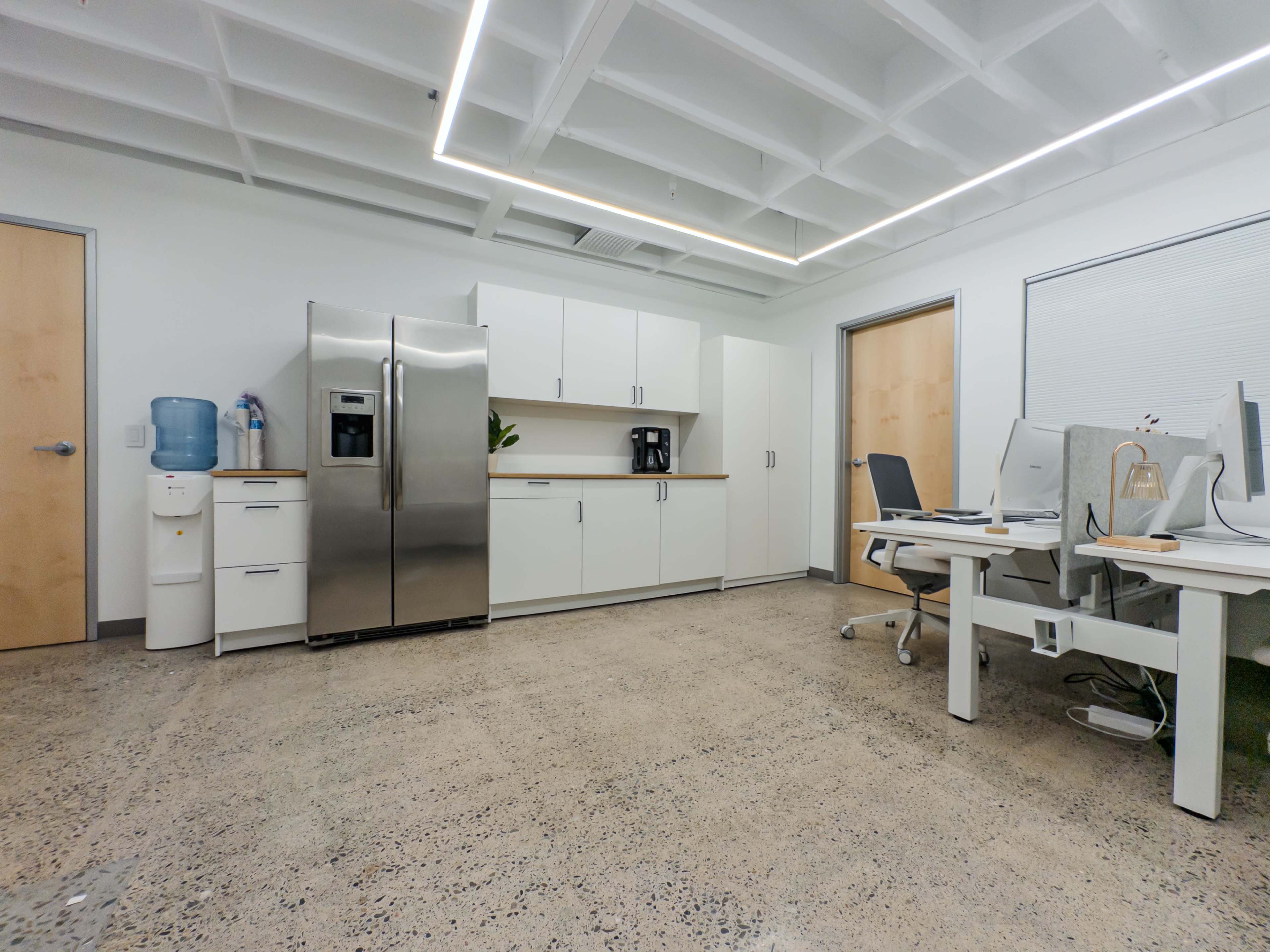 The image shows a modern office space featuring a stainless steel refrigerator, a coffee machine, and two workstations with computer monitors, all set against a concrete floor.