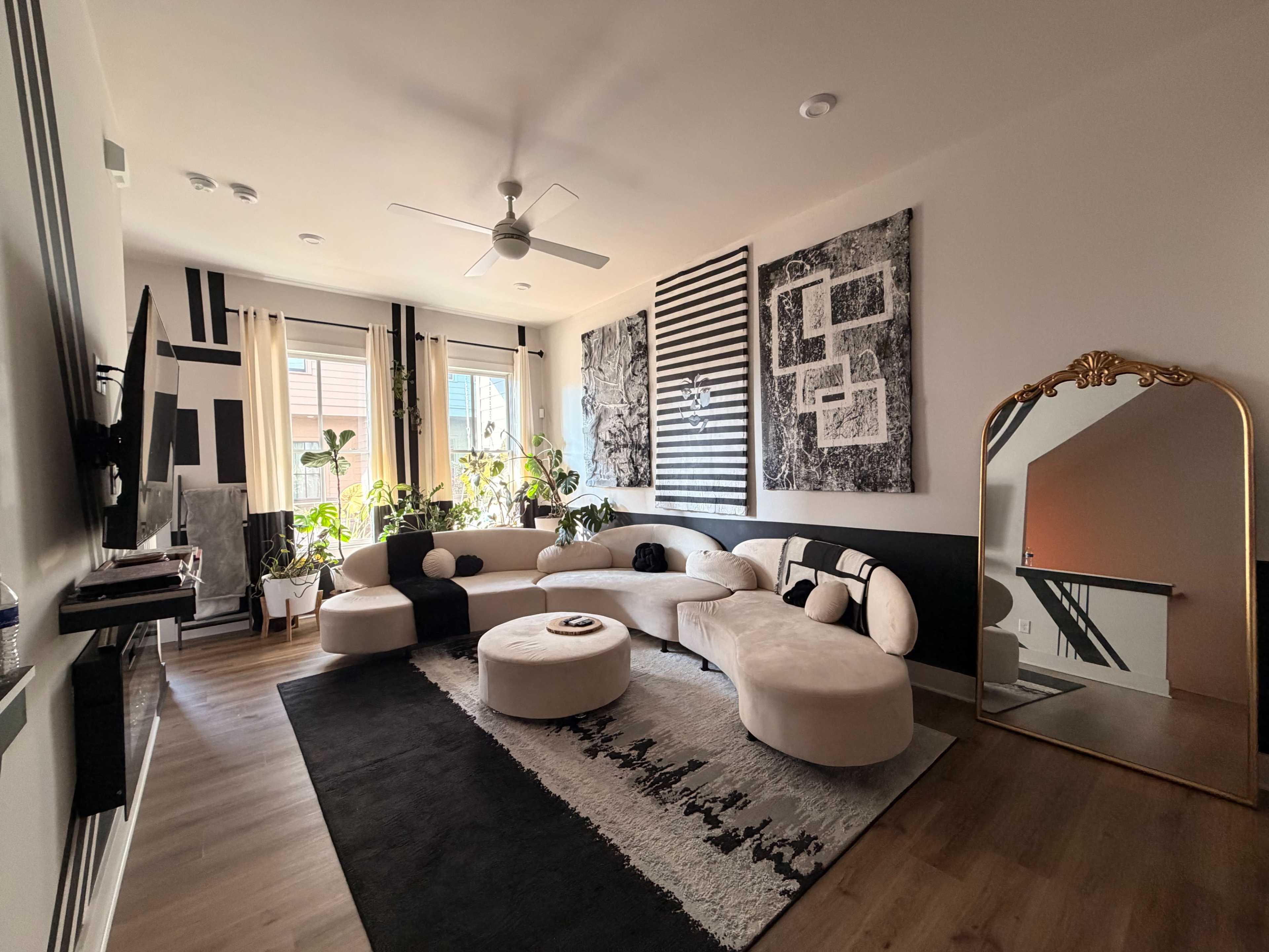 The living room features a modern sectional sofa, decorative wall art, large windows with natural light, and a gold-framed mirror.