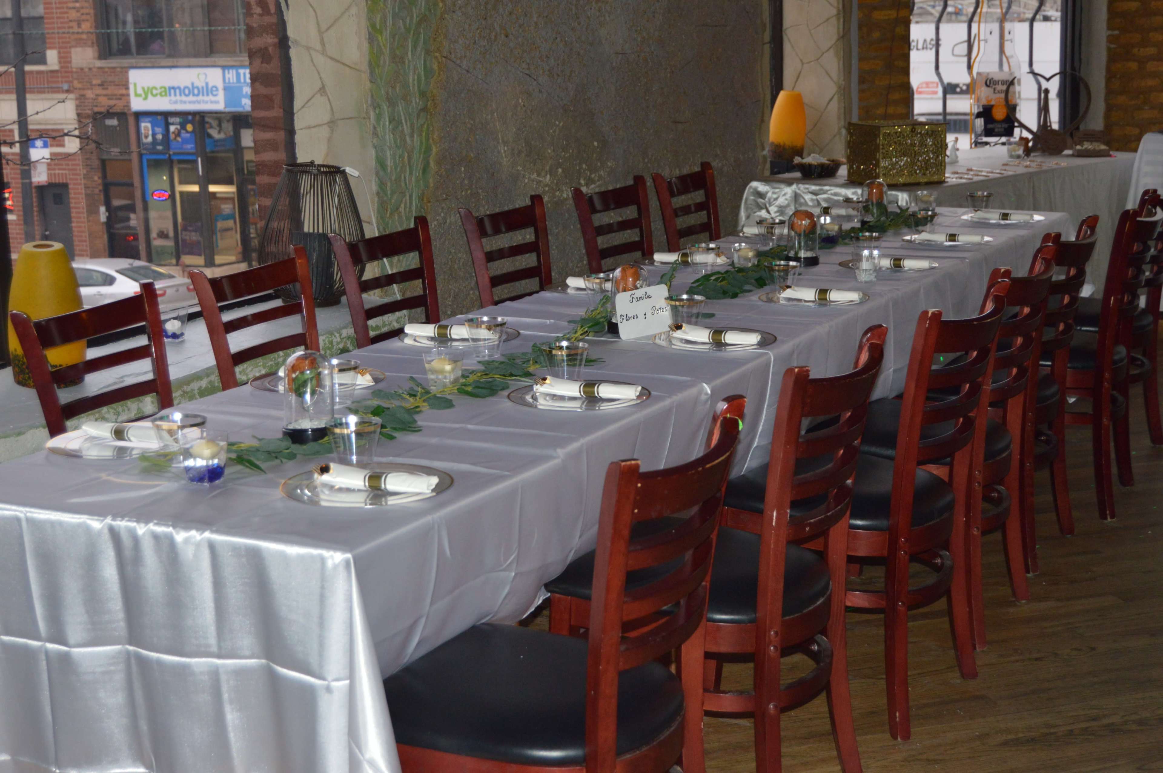 A long dining table is set with plates, utensils, and glass candles, decorated with greenery, in a restaurant setting.