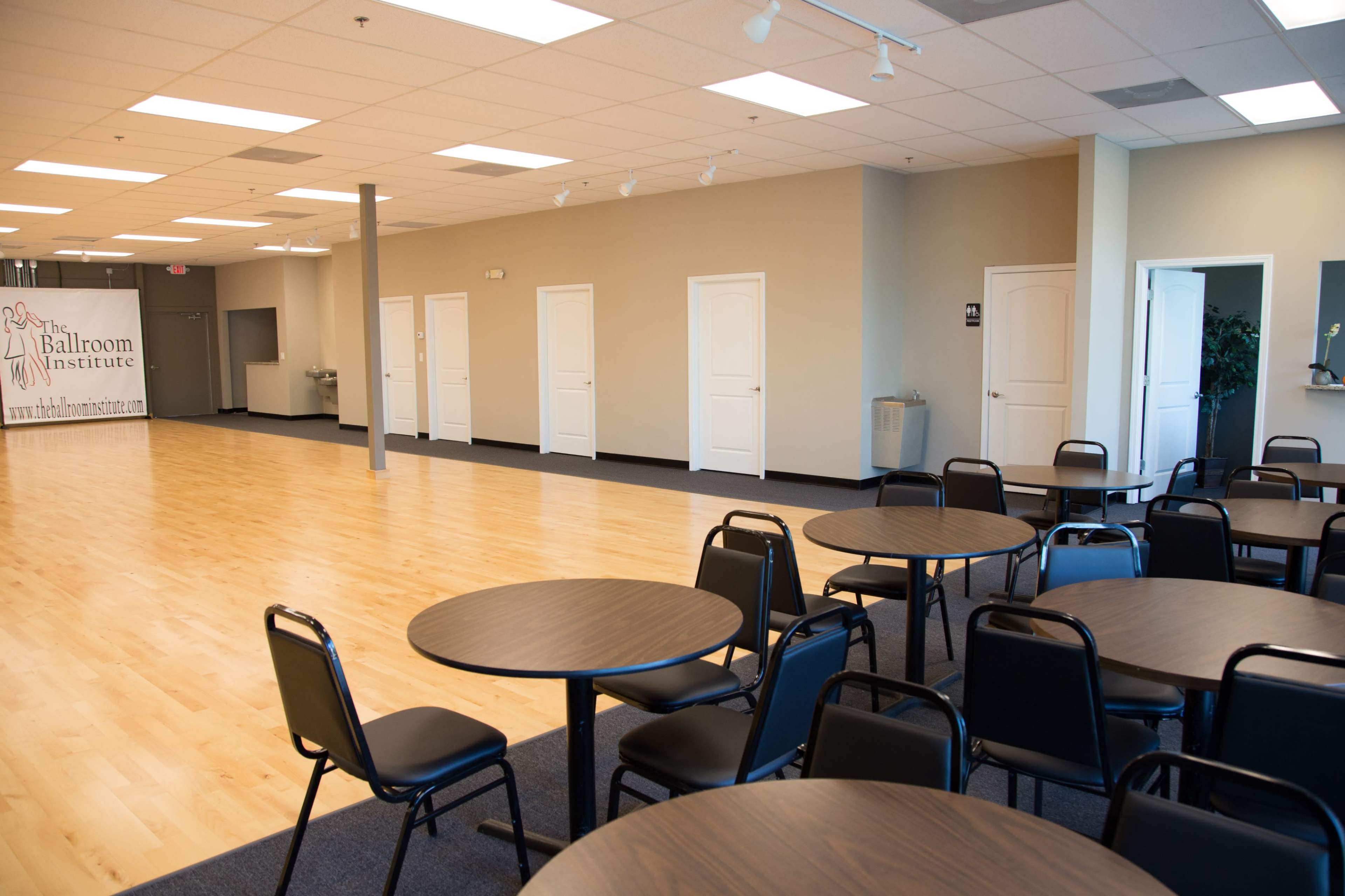 The image shows a spacious, well-lit room with several round tables and black chairs arranged in the foreground, while multiple closed doors line the walls in the background.