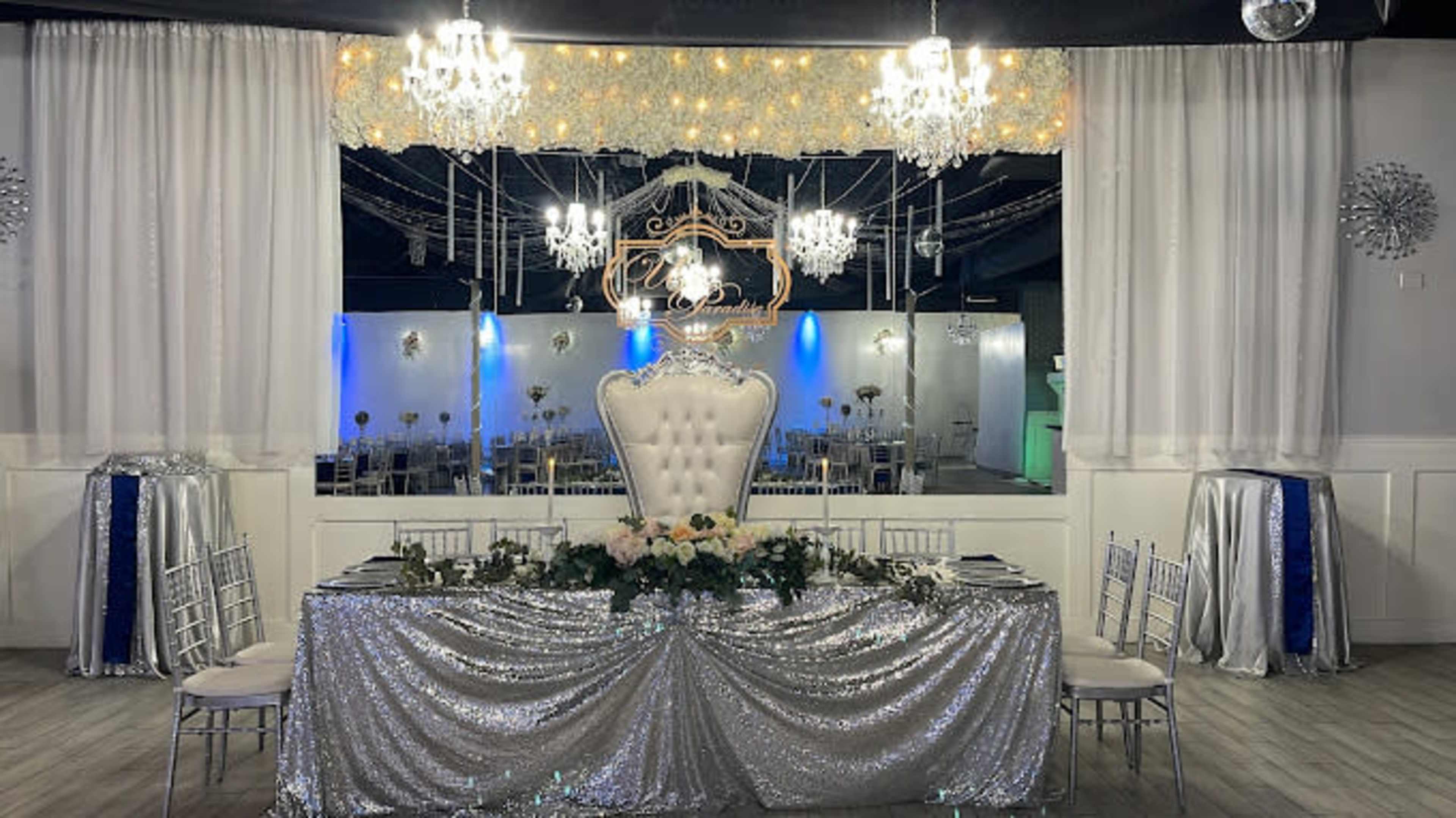 A formal dining setup features a sparkling silver tablecloth and elegant white chairs, adorned with floral arrangements and chandeliers in a decorated event space.