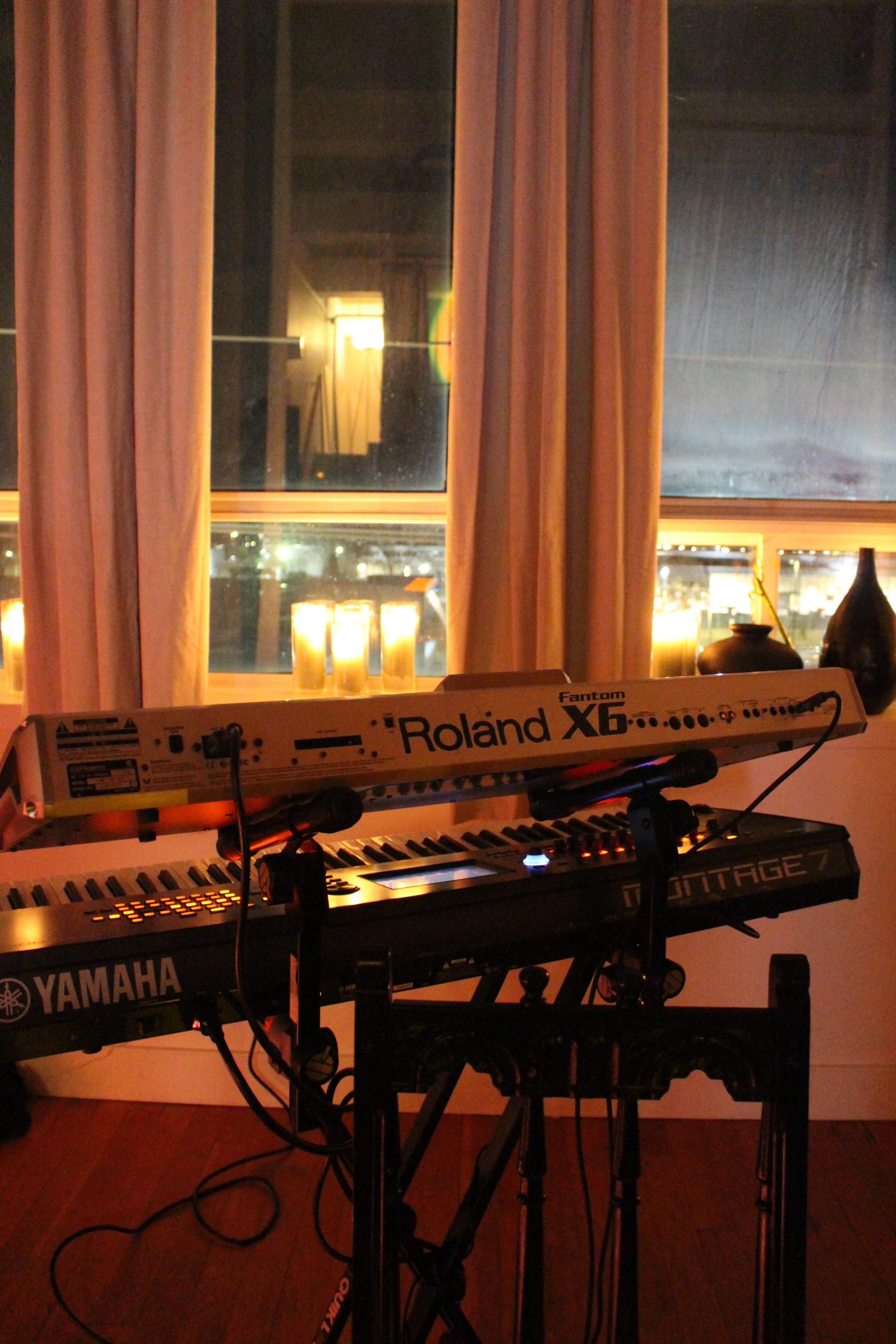 A Yamaha and a Roland keyboard are positioned on a stand in front of a window, with soft candlelight illuminating the scene.