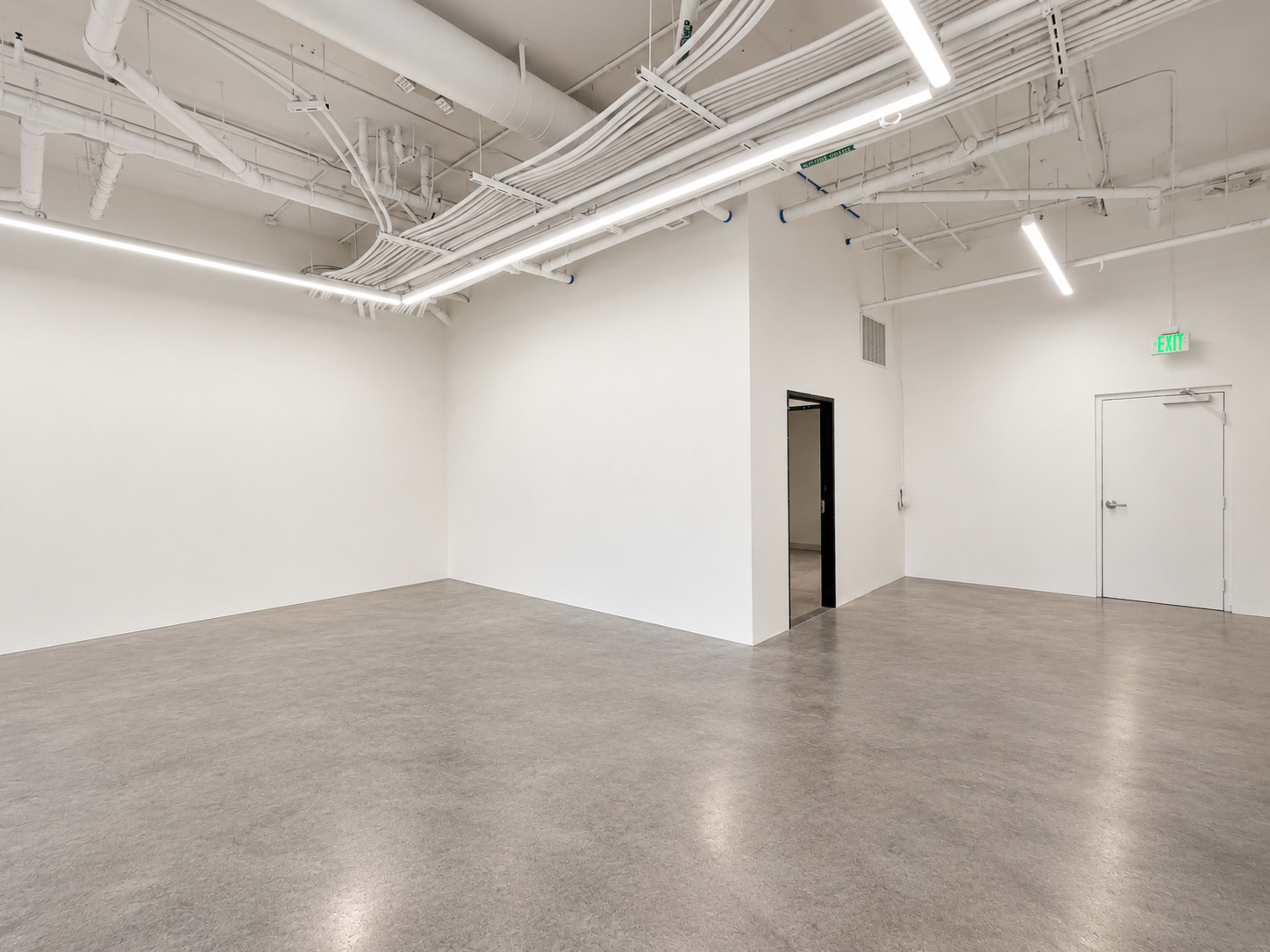 The image shows a spacious, empty interior with smooth, light-colored walls and a polished floor, featuring various overhead pipes and fluorescent lighting.