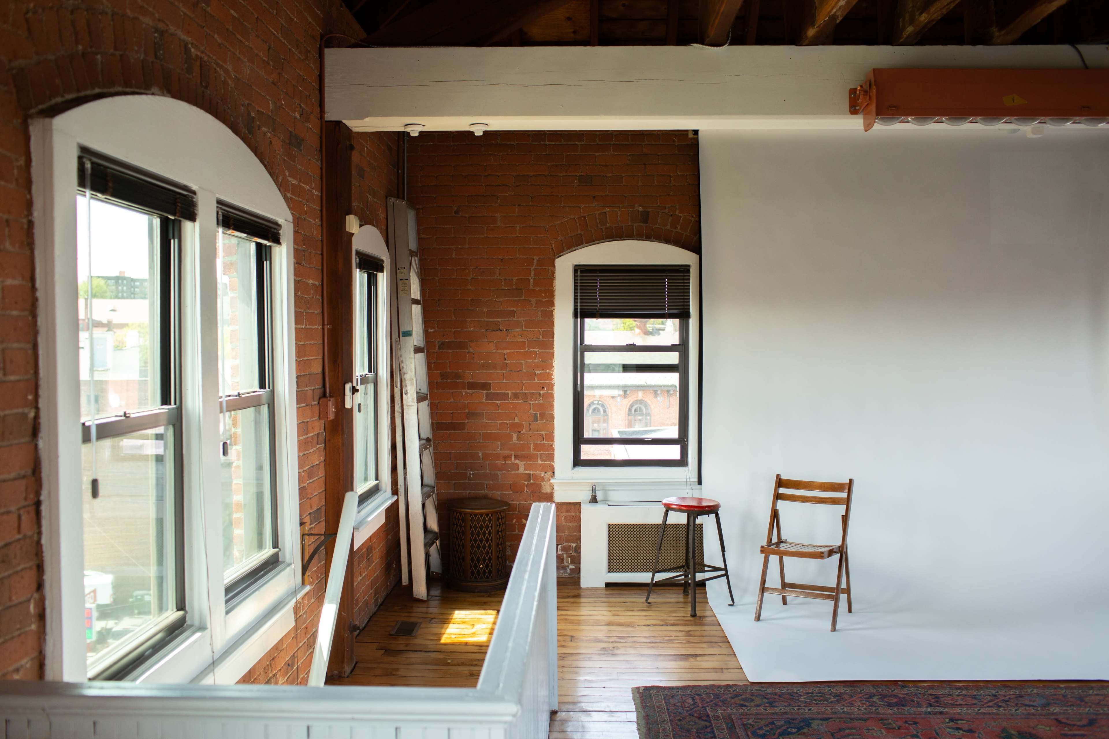 A spacious room with exposed brick walls features a ladder, a chair, and a plain backdrop against the far wall.