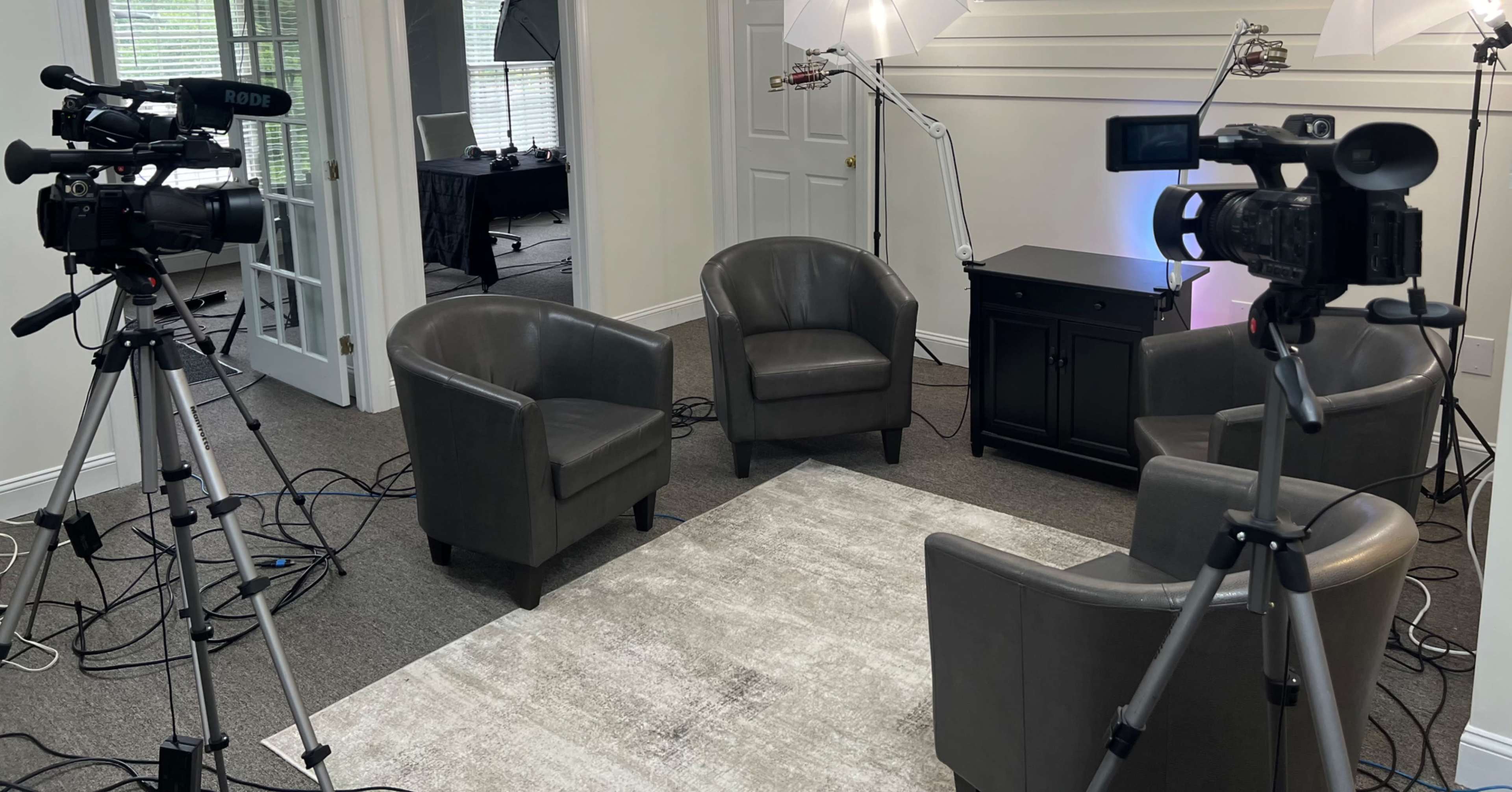 A small interview setup featuring three upholstered chairs arranged around a coffee table, with video cameras and lighting equipment positioned nearby.