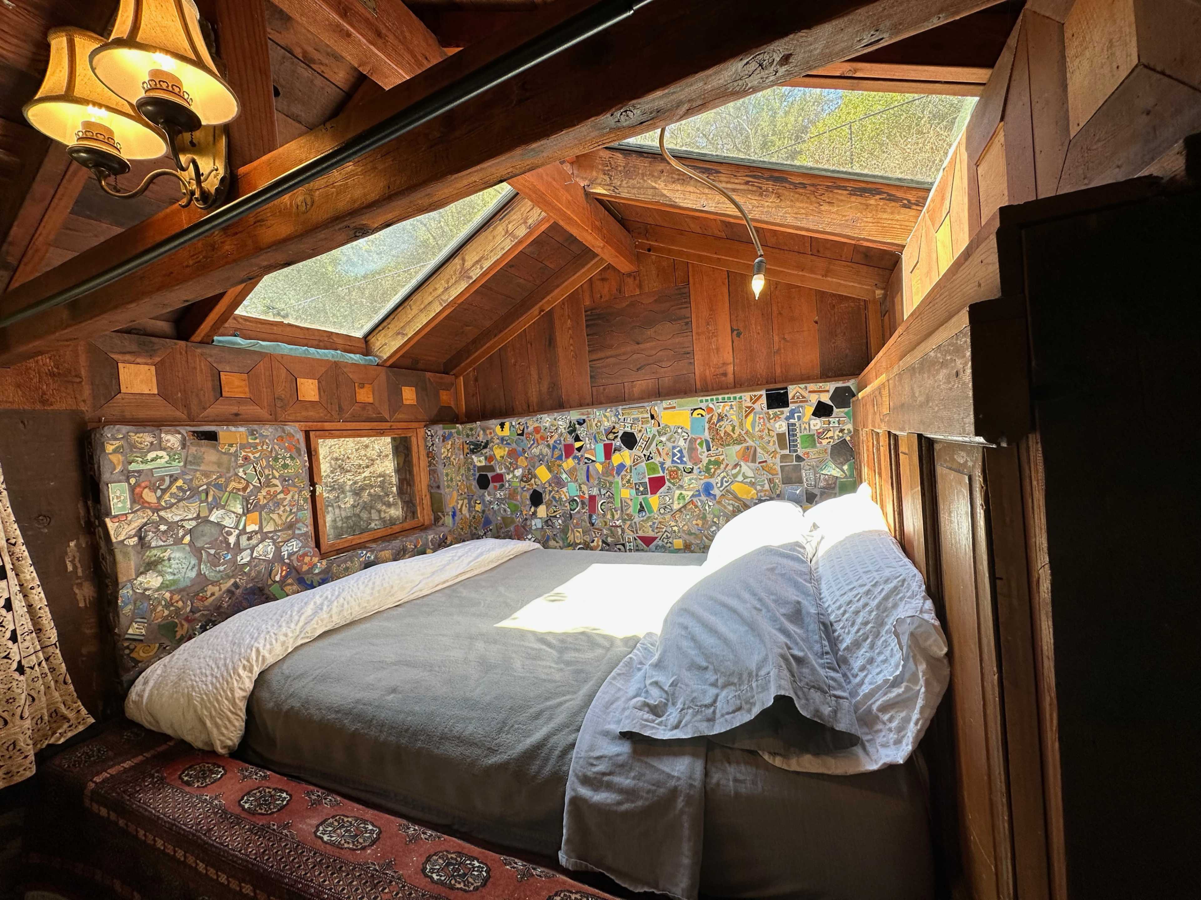 A rustic bedroom with a skylight, a bed with gray linens, and a colorful mosaic wall.