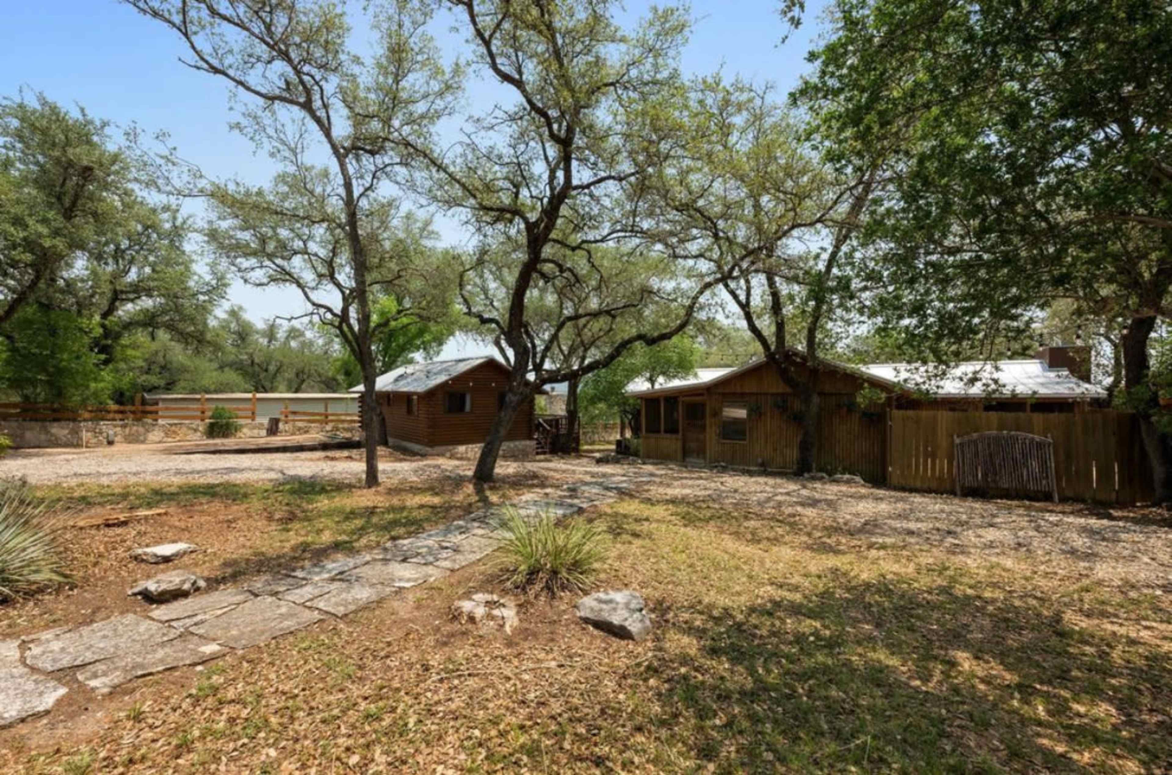The image shows a wooded area with two rustic cabins surrounded by trees and a stone pathway leading through a natural landscape.