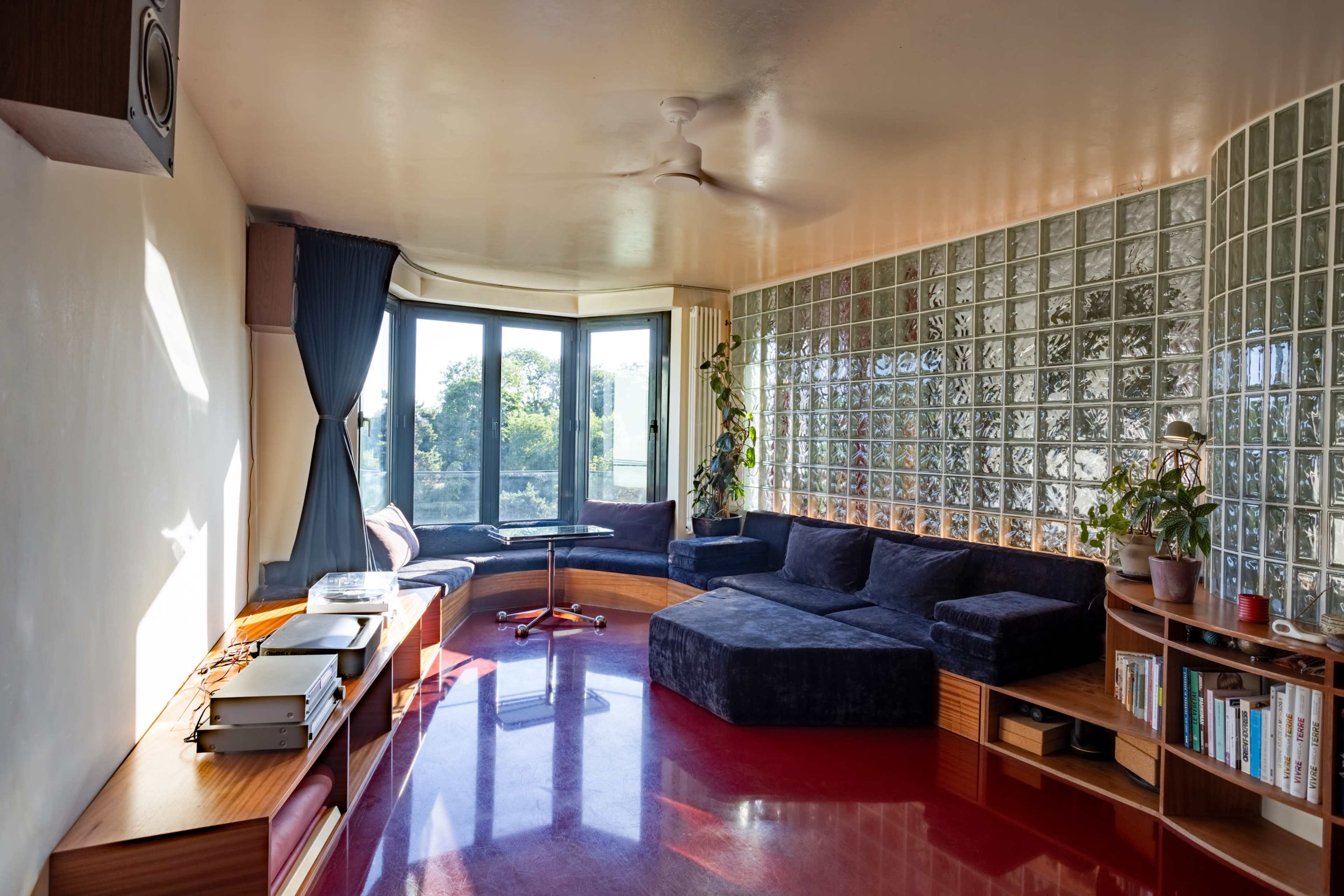 The image shows a modern living room with a curved sectional sofa, a glass block wall, and large windows overlooking a green landscape.