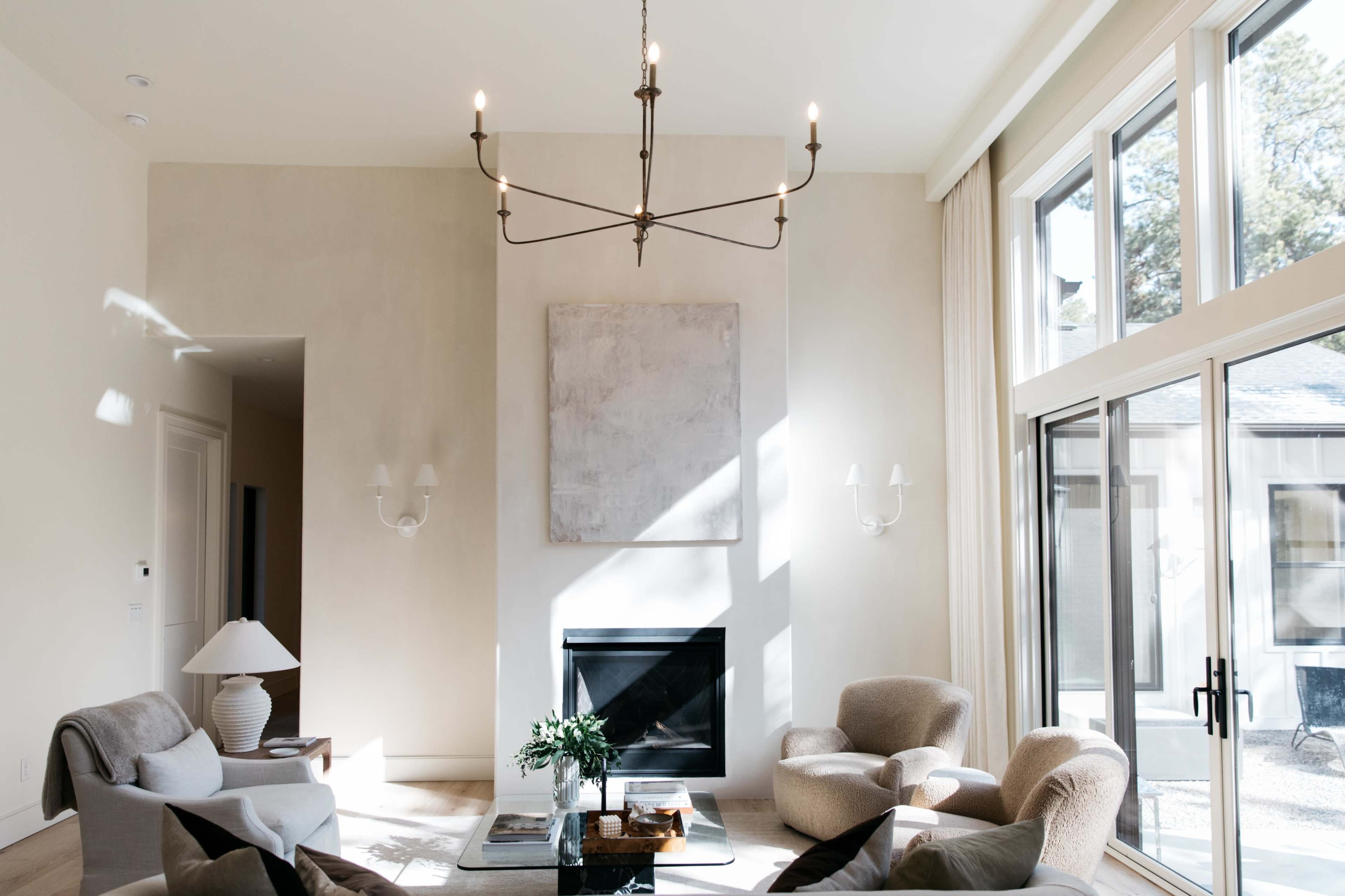 A spacious living room features a modern chandelier, a central fireplace, and large windows allowing natural light to fill the space.