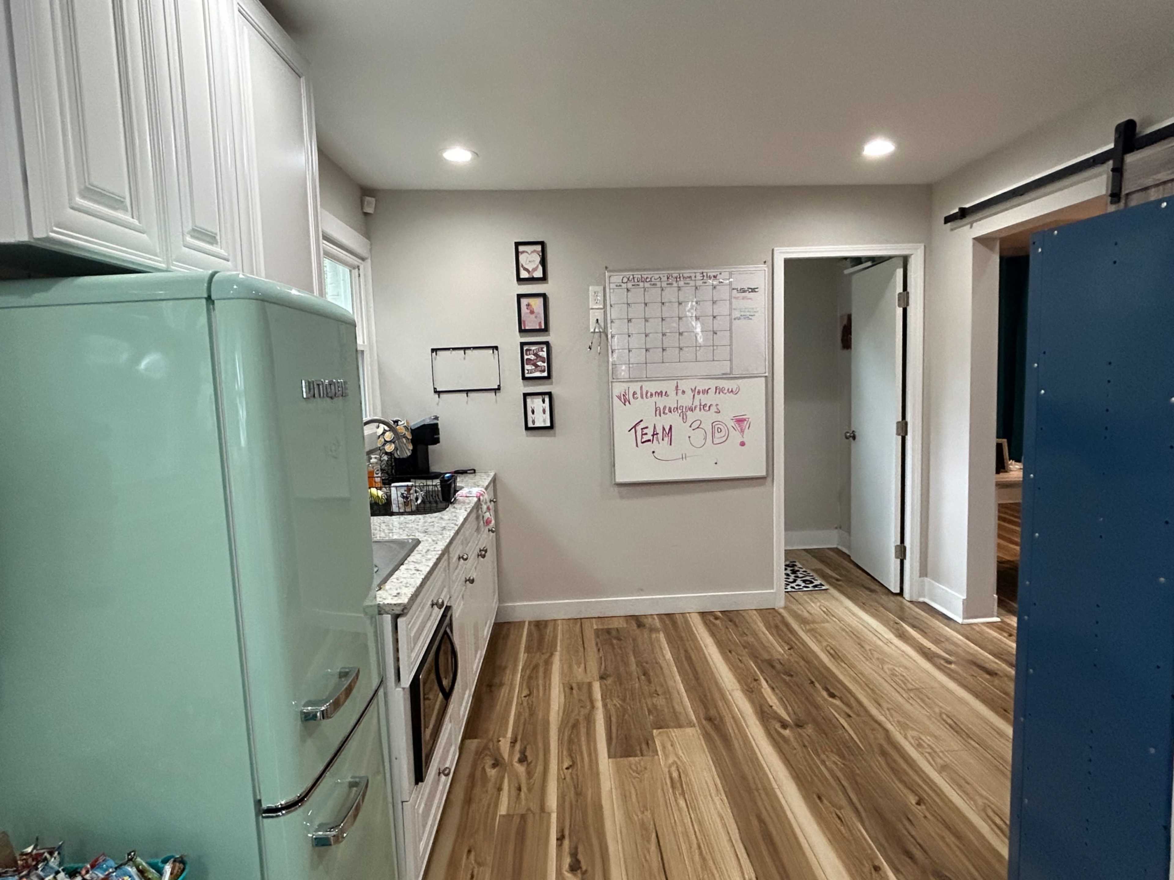 The image shows a modern kitchen with light-colored cabinets, a mint green refrigerator, a whiteboard on the wall, and wooden flooring.