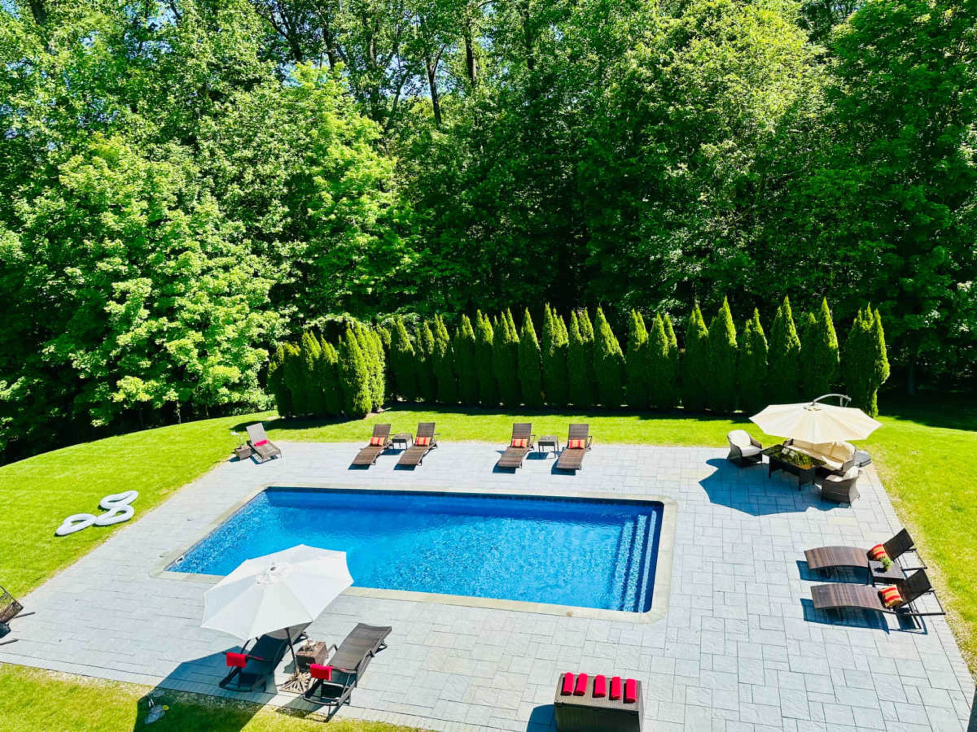 The image shows a rectangular swimming pool surrounded by lounge chairs and two umbrellas, set in a manicured grass area bordered by tall green trees.