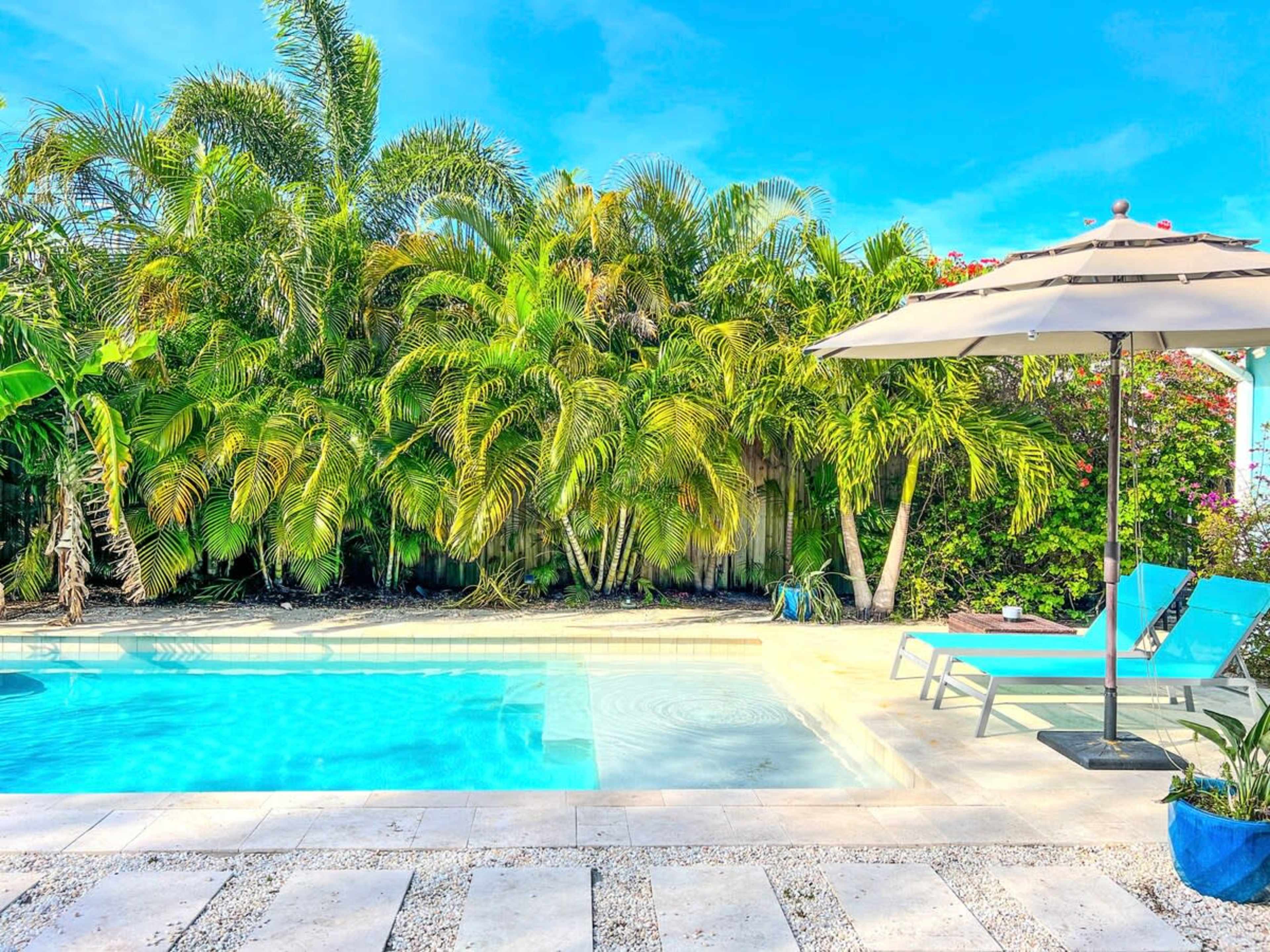 A clear swimming pool is surrounded by lush palm trees and two blue lounge chairs next to a beige umbrella.