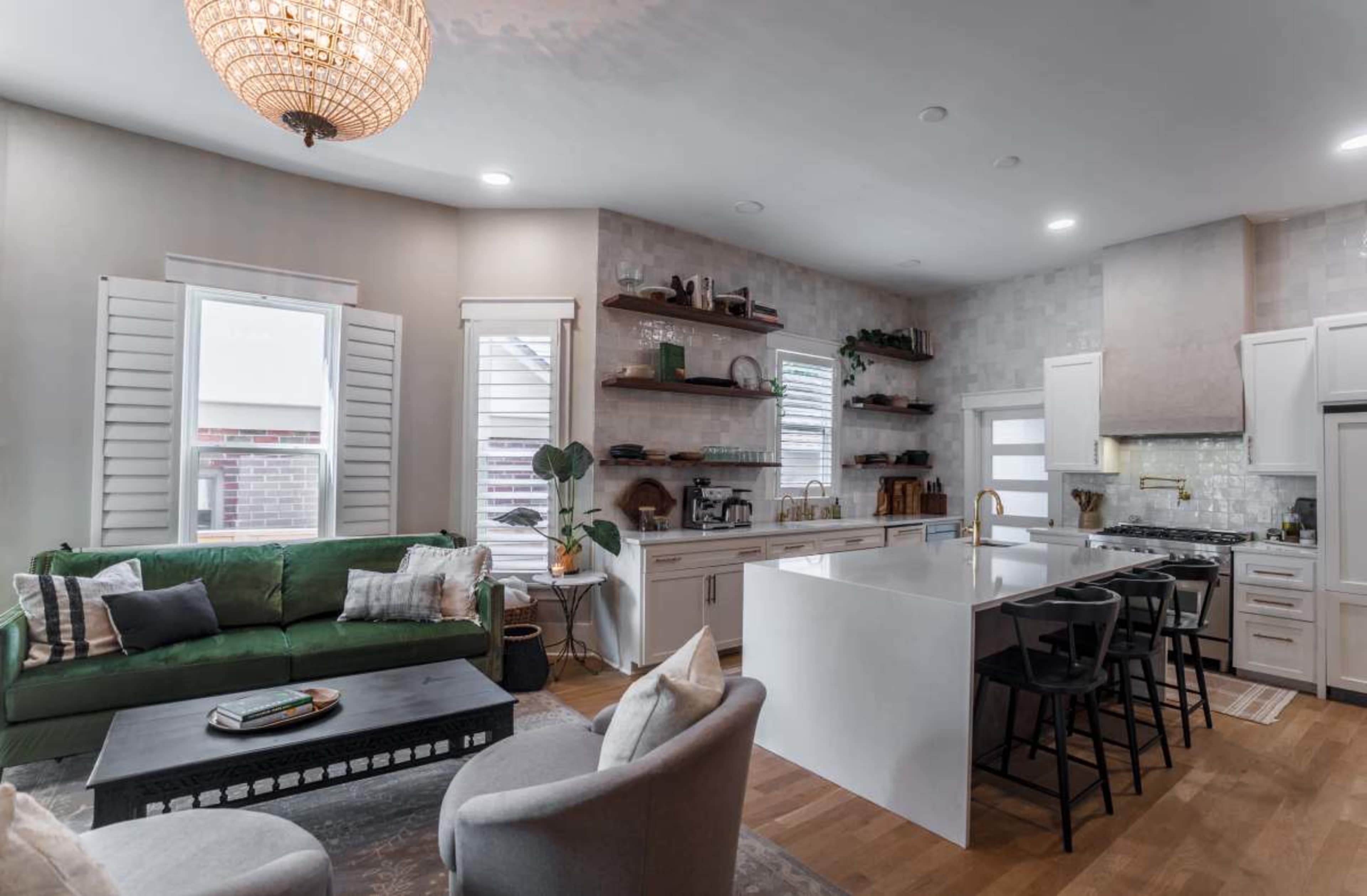 The image shows a modern kitchen and living area featuring a white kitchen island, a green sofa with decorative pillows, and large windows allowing natural light.
