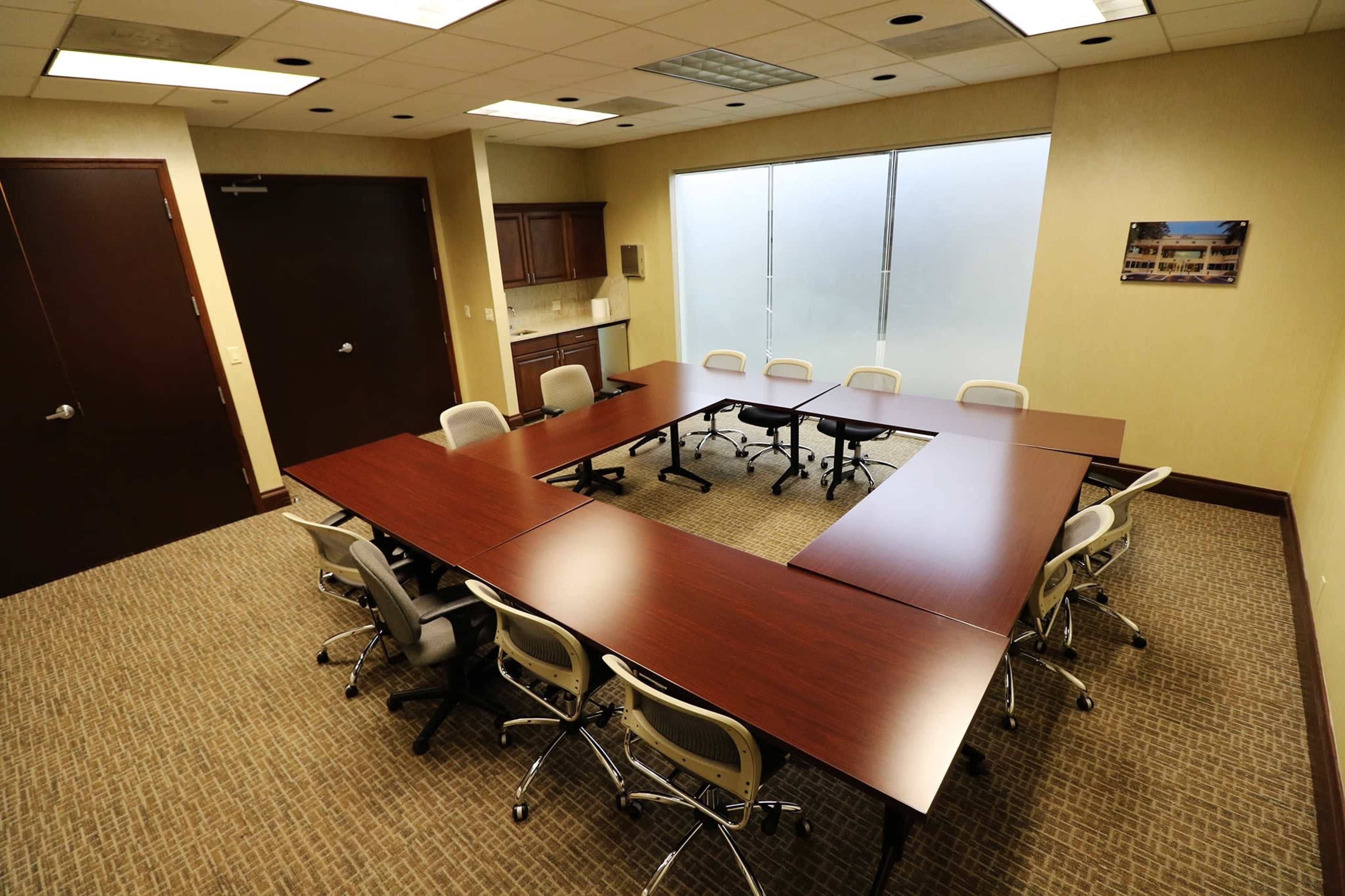 A rectangular conference room features a large, U-shaped table surrounded by office chairs, with a kitchenette and a frosted glass window in the background.