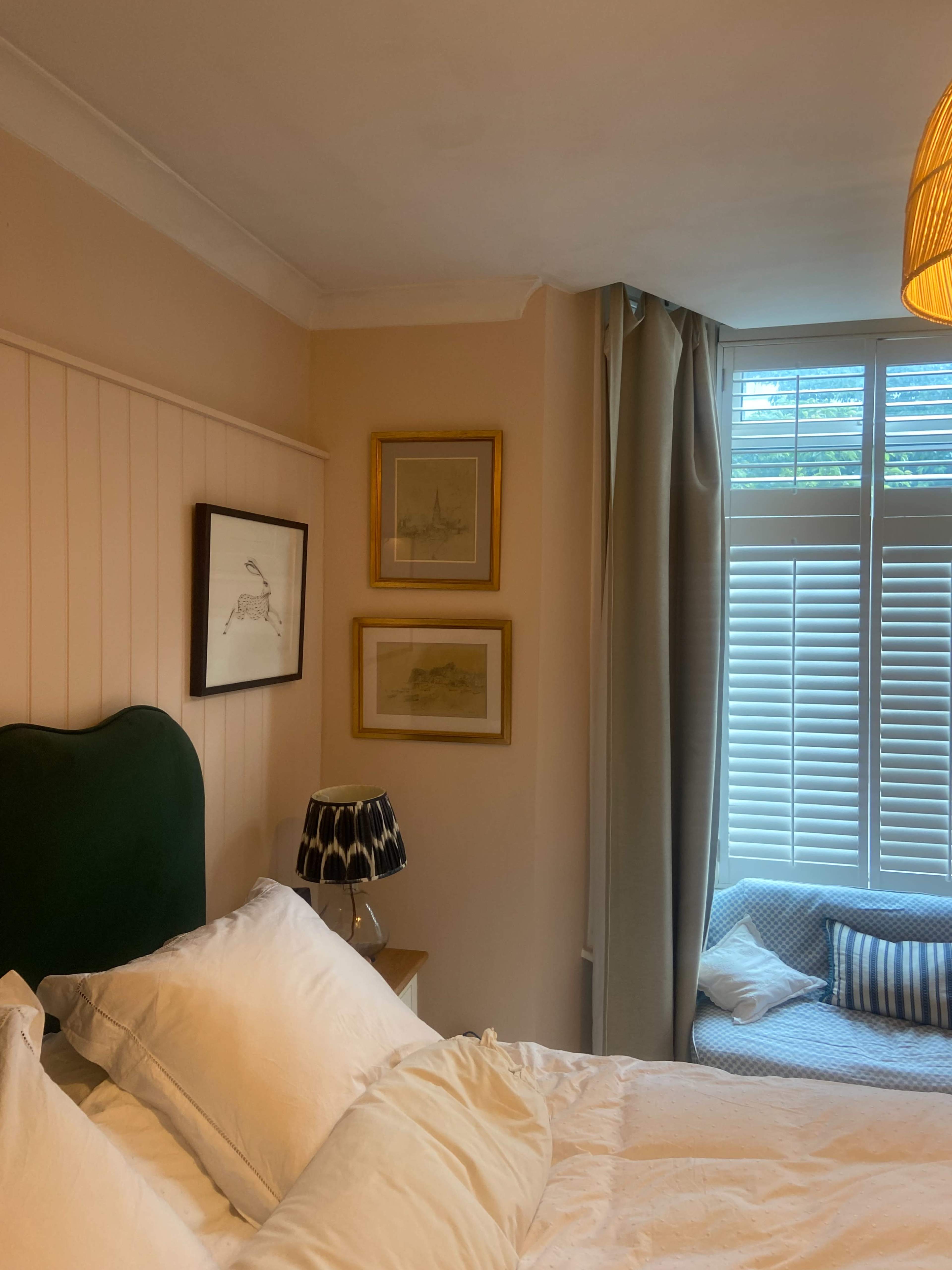 The image shows a cozy bedroom with a green upholstered headboard, framed artwork on the walls, a lamp on a nightstand, and a window with light-colored curtains and a window seat.