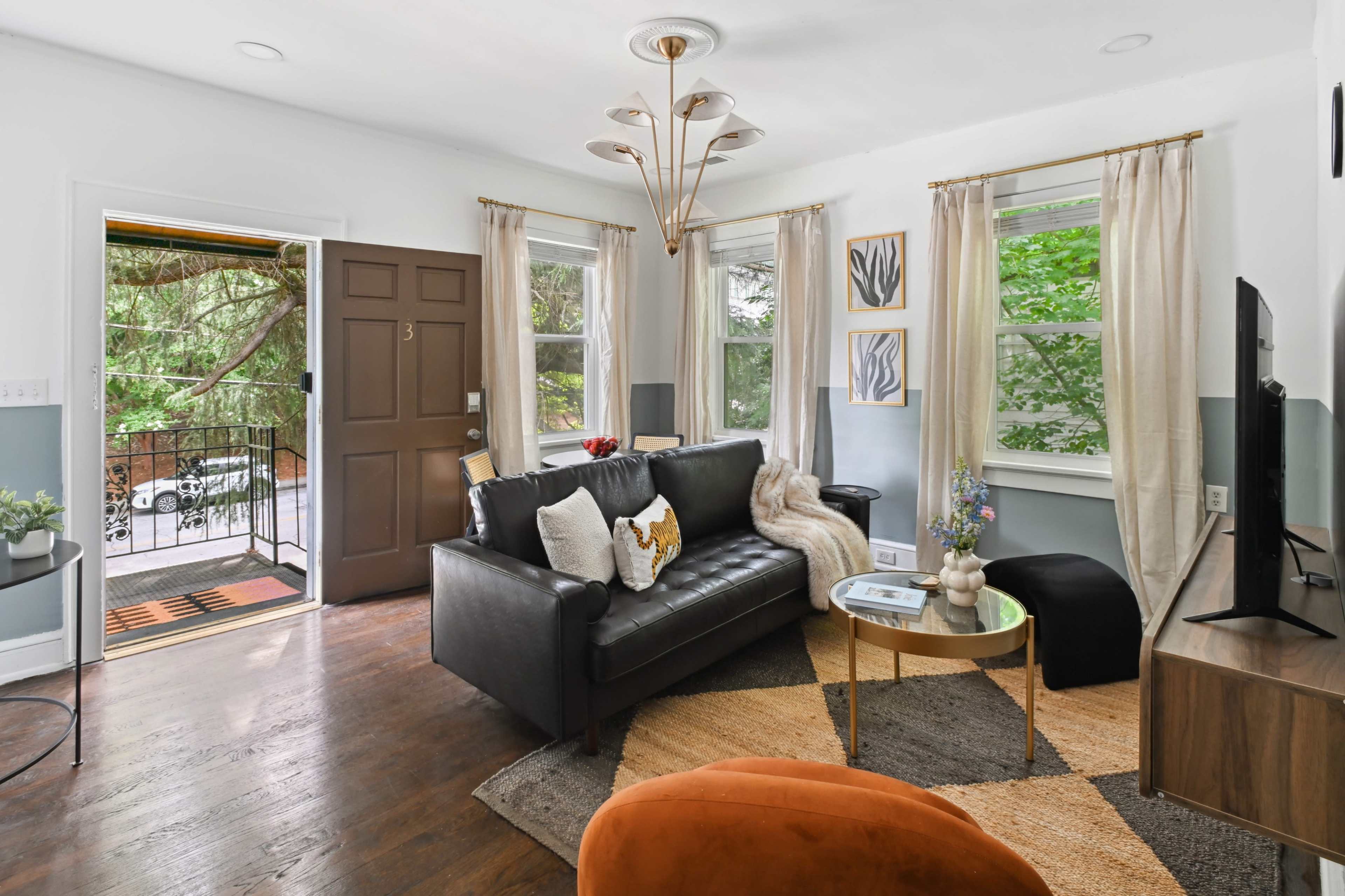 The living room features a black sofa, a round gold coffee table, and large windows with sheer curtains, opening to a view of greenery outside.
