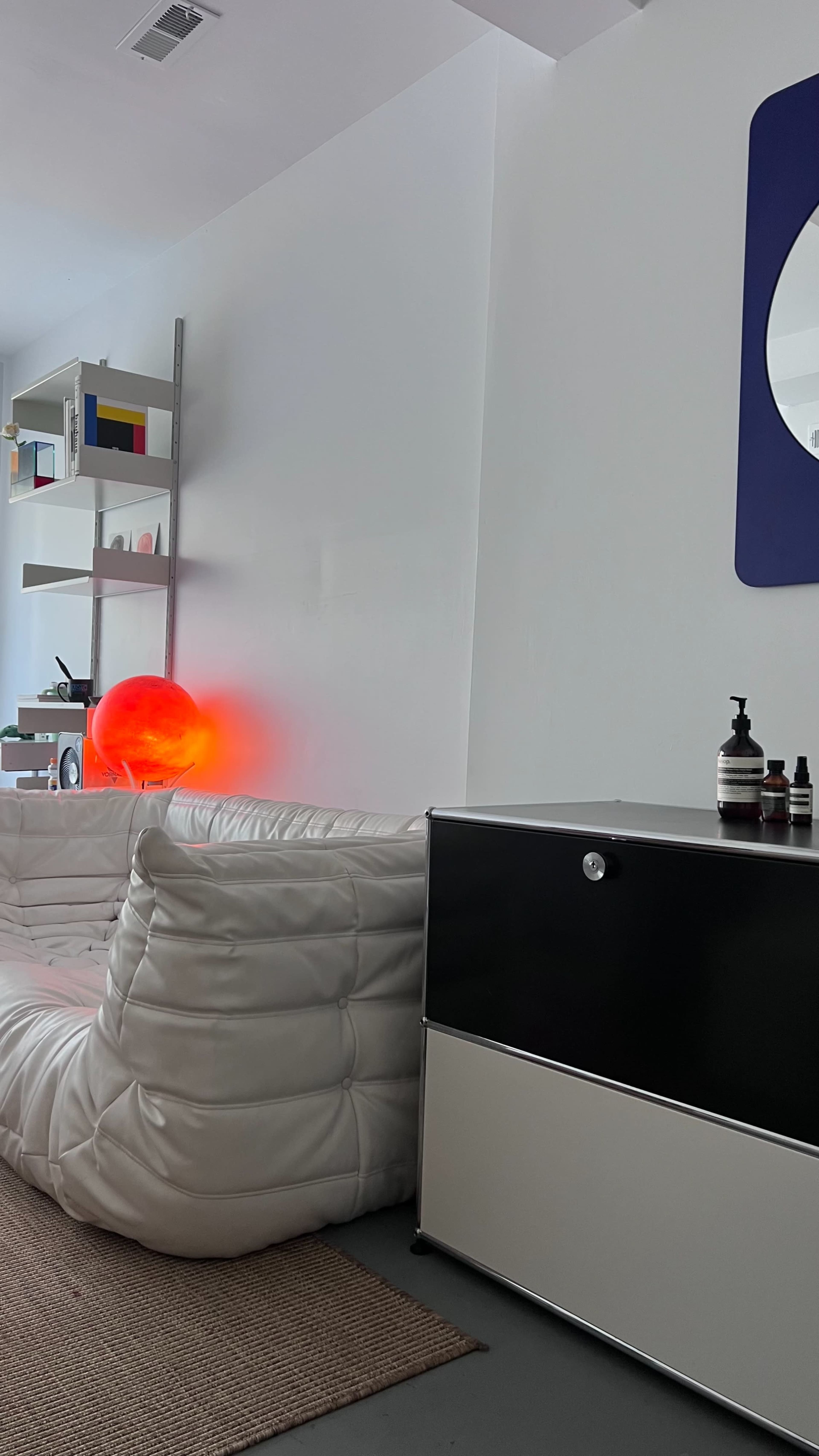 A contemporary living space features a white modular sofa, a round mirror, a black and white chest of drawers, and a glowing red orb on the floor.