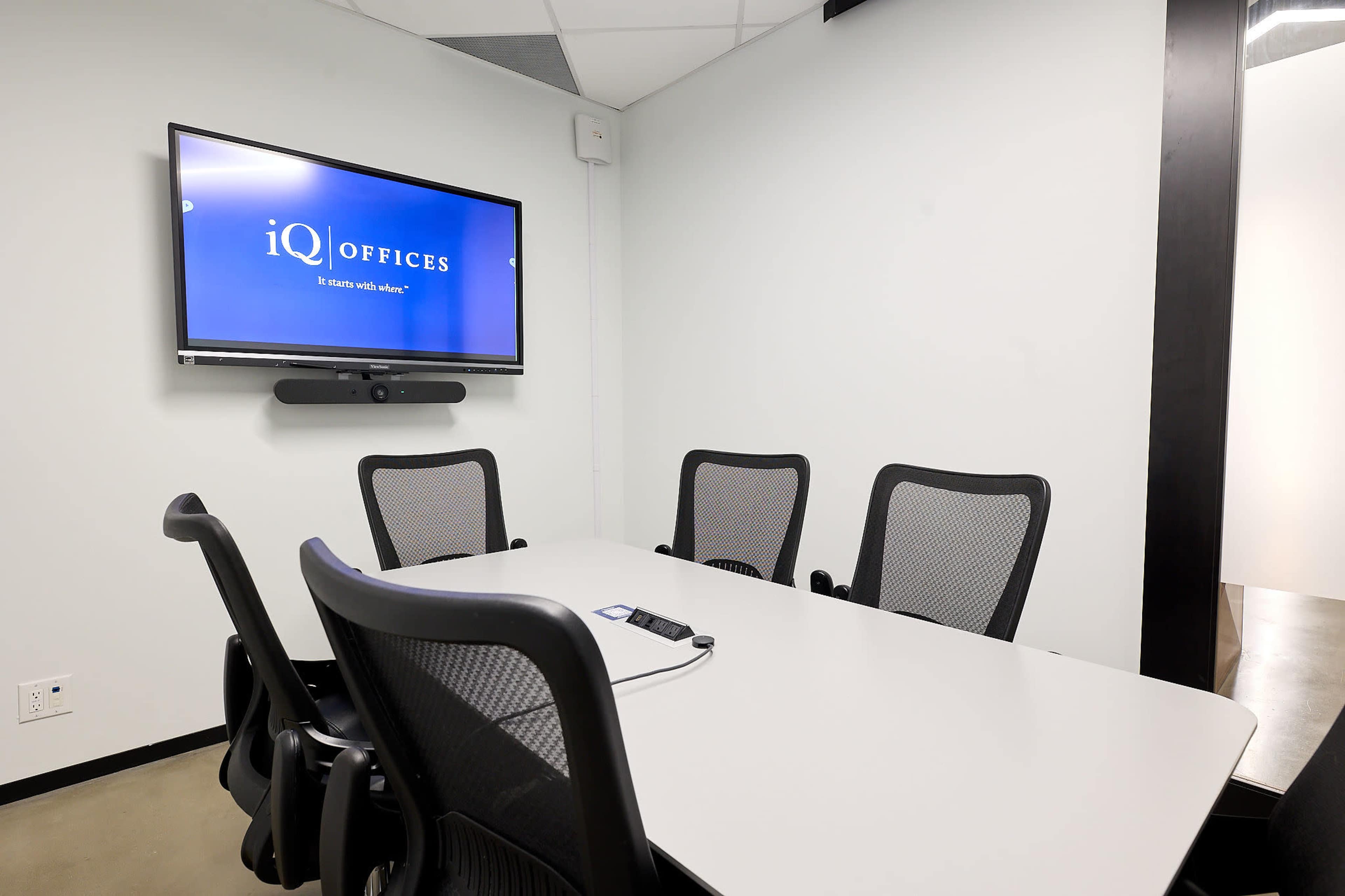 The image shows a small meeting room with a rectangular table and four ergonomic chairs, featuring a wall-mounted television displaying the logo of iQ Offices.