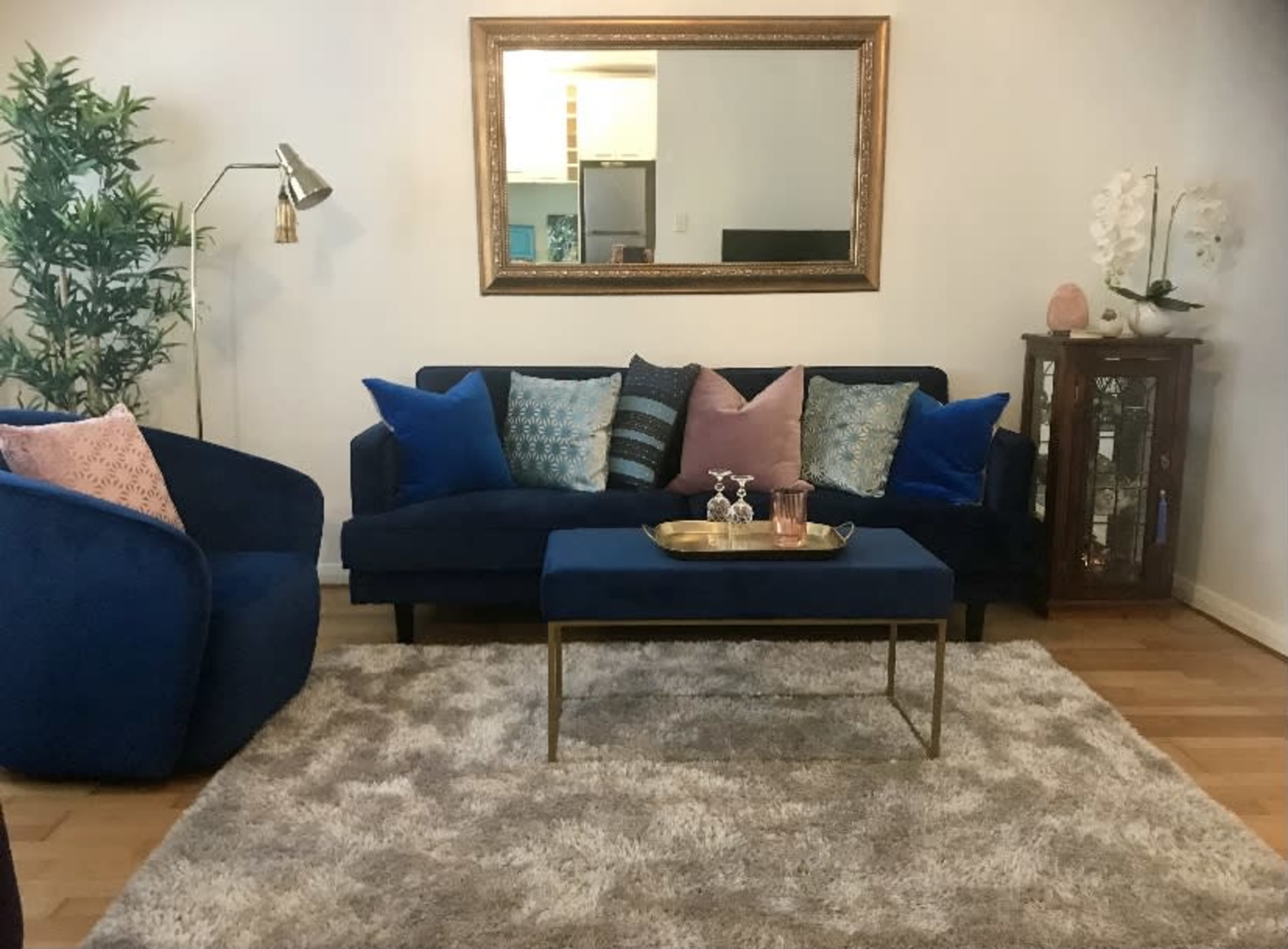 A living room features a navy blue sofa with decorative pillows, a small coffee table with a tray, and a large mirror above it, all arranged on a soft rug.