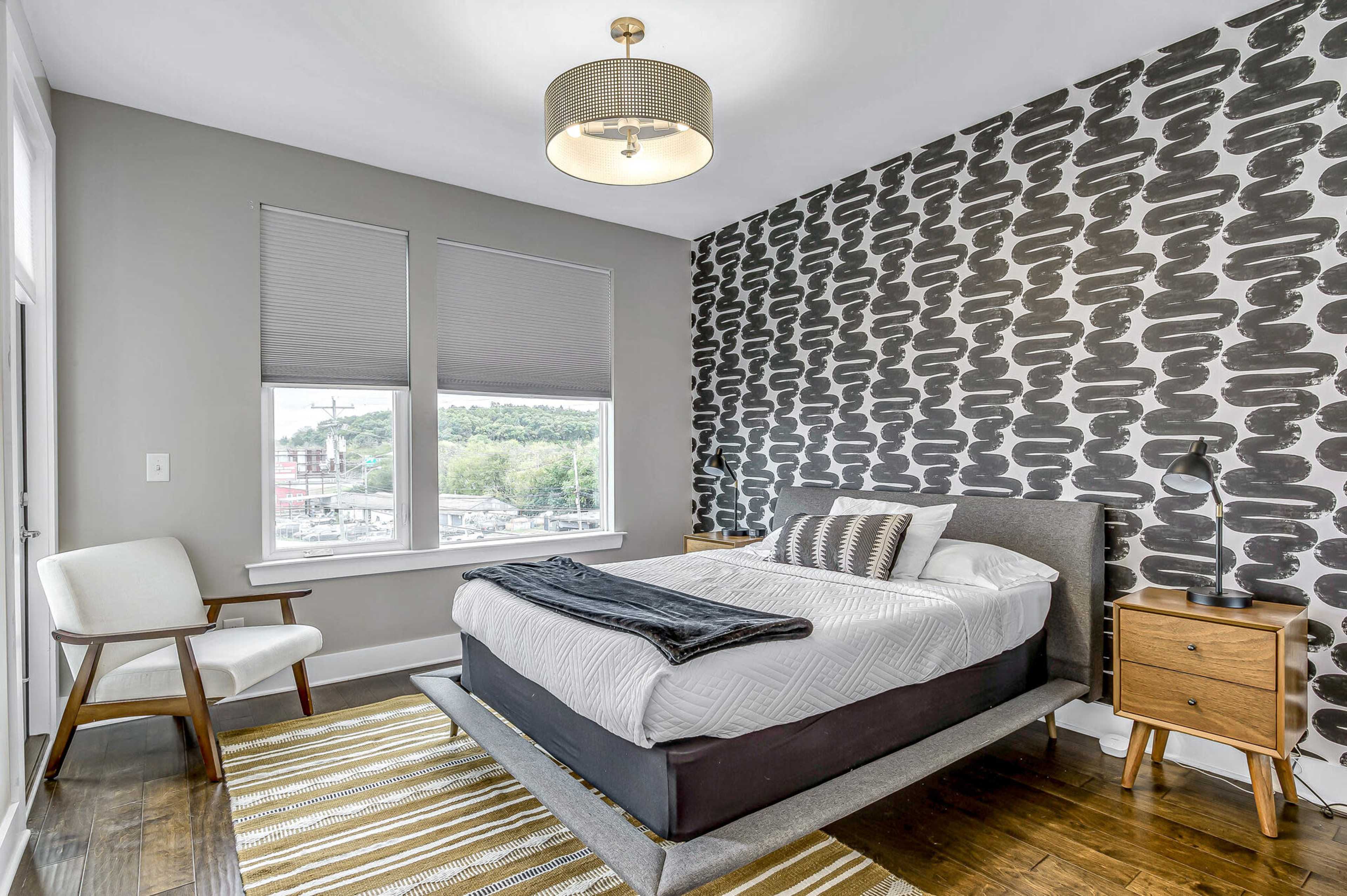 The image shows a modern bedroom with a gray-patterned accent wall, a bed with a black and white throw blanket, a stylish chair, and a bedside table with a lamp.