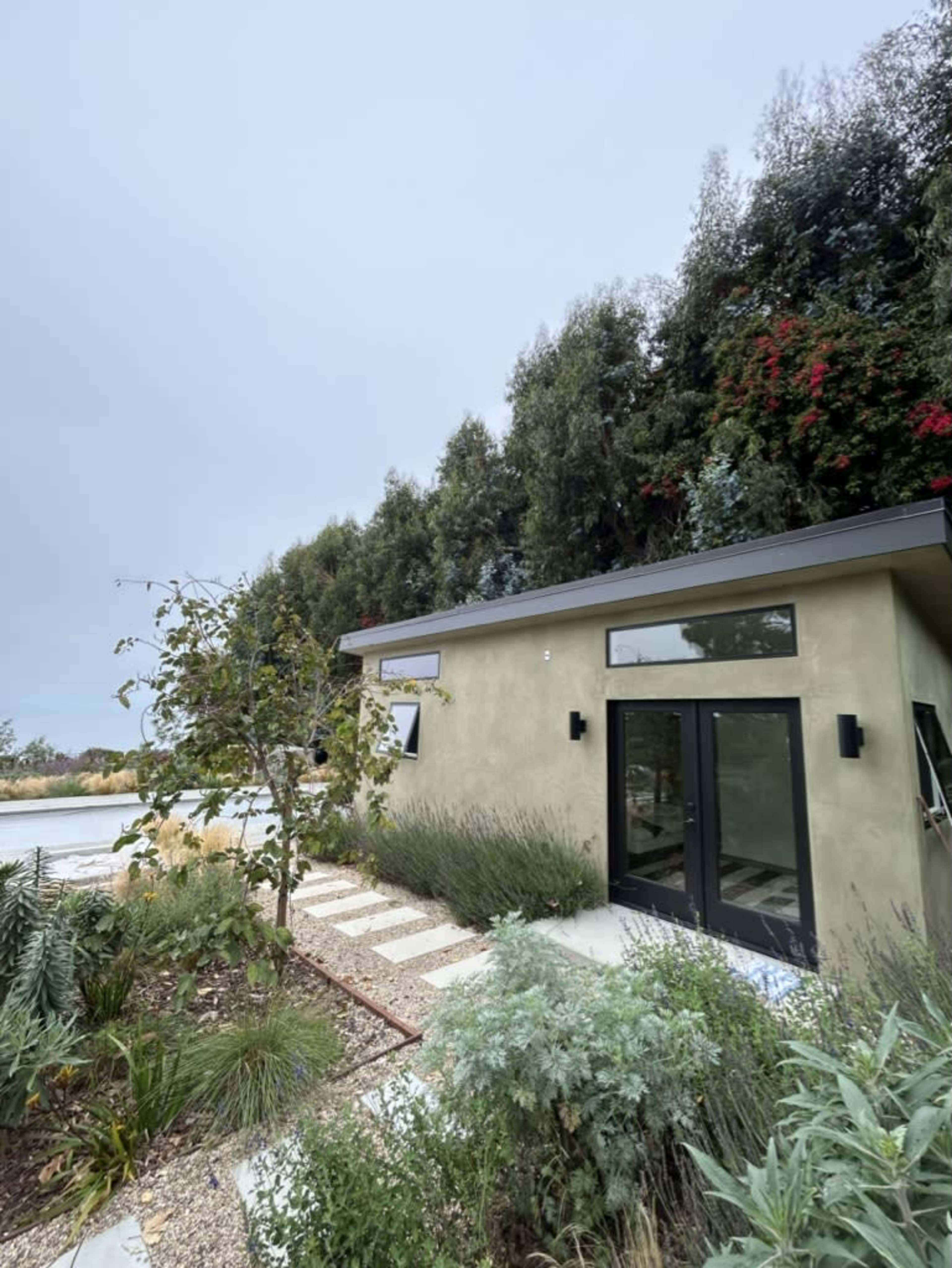 A small, modern house with large windows and a stone pathway is surrounded by various plants and trees.