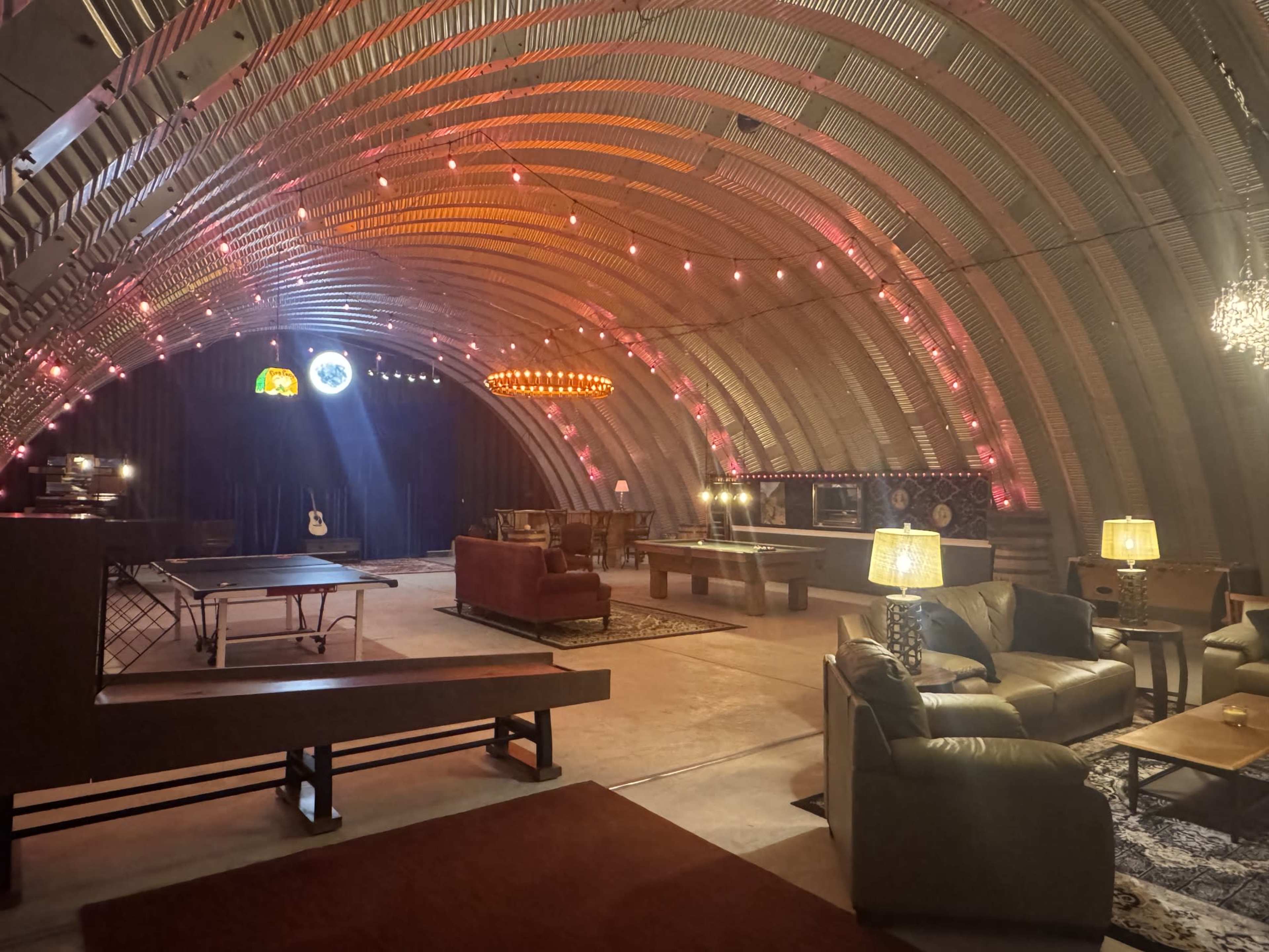 The image shows a spacious, industrial-style room with a vaulted ceiling, featuring lounge seating, a ping pong table, and illuminated decorations.