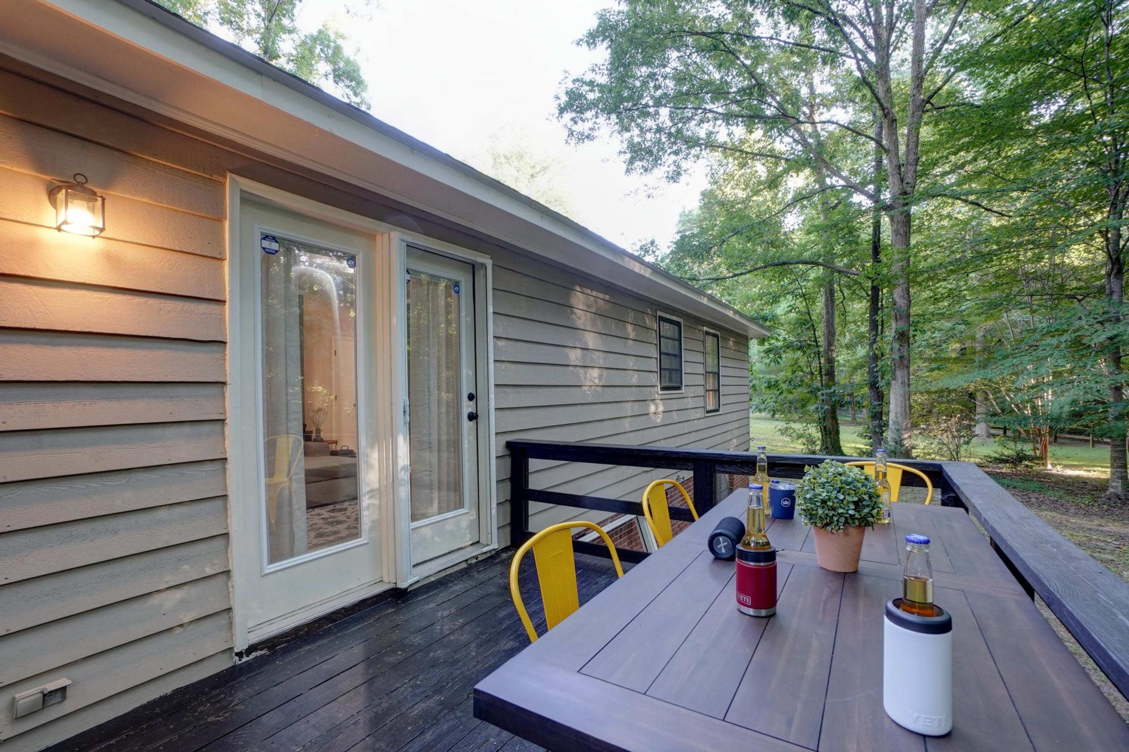 A wooden deck with a table and chairs overlooks a backyard surrounded by trees, with a set of glass doors leading into the house.
