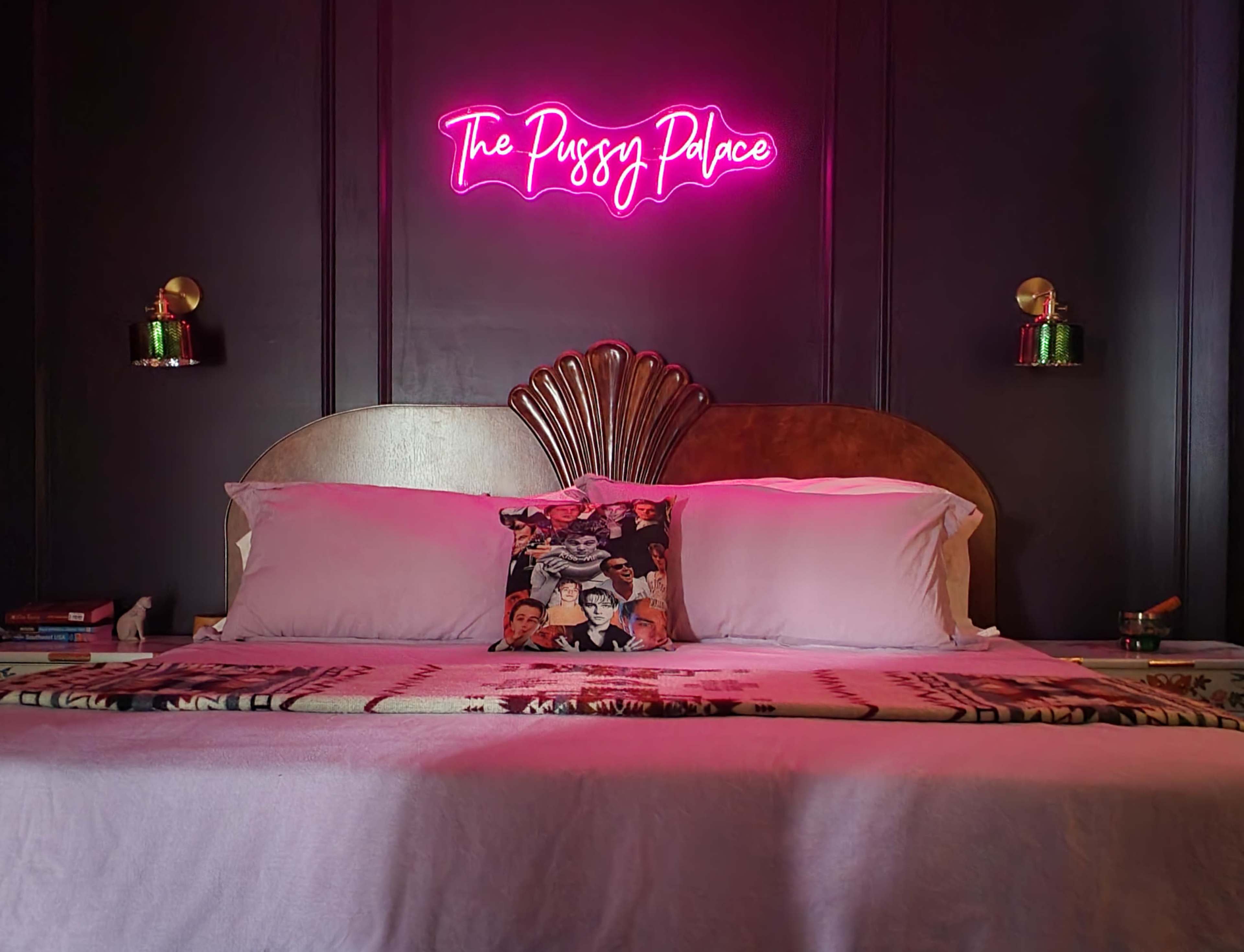 A bed with a decorative headboard is centered against a dark wall, featuring a neon sign reading "The Pussy Palace" above it.