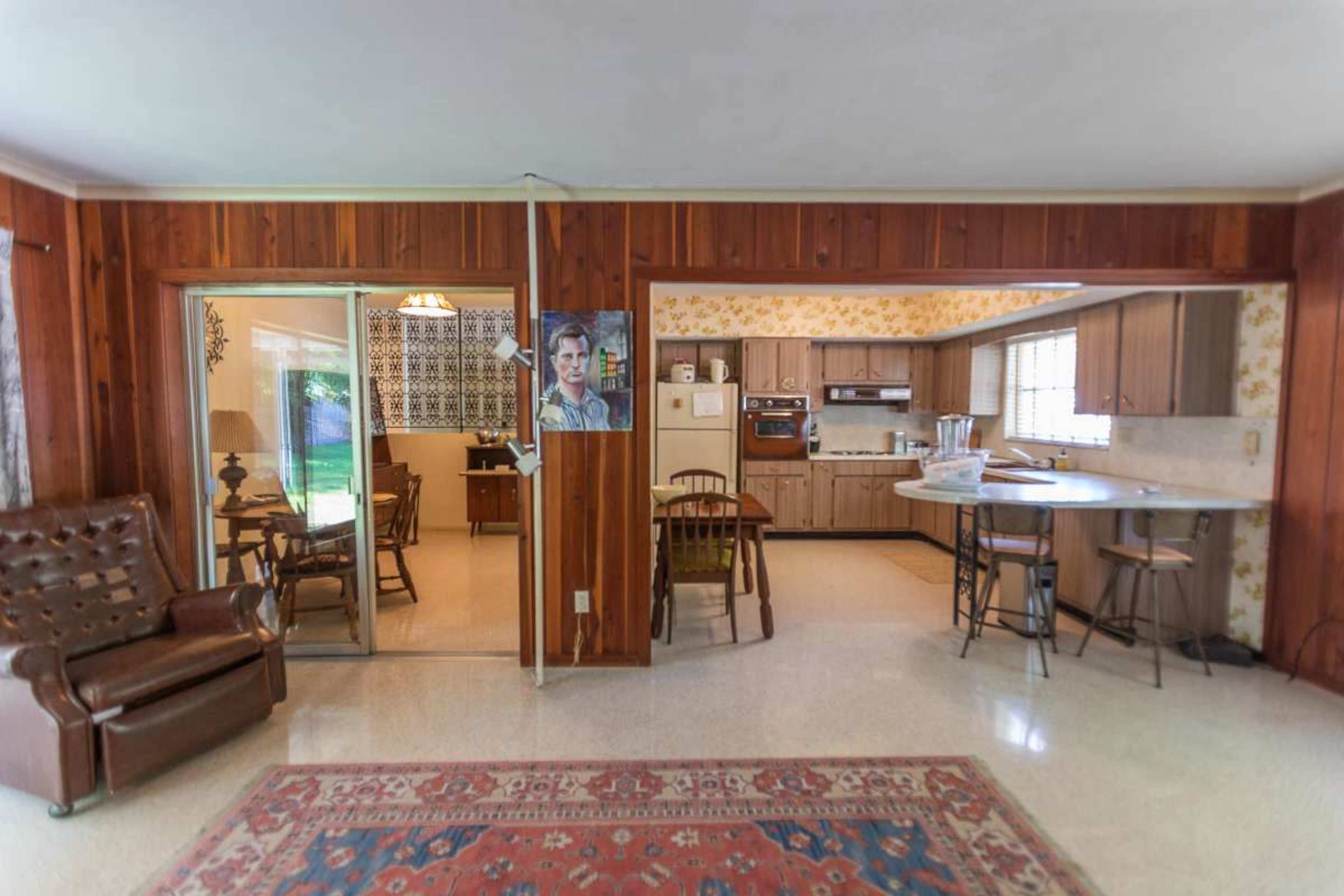 The image shows a living area that opens into a kitchen, featuring wooden paneling, a patterned wallpaper, and a vintage-style rug.
