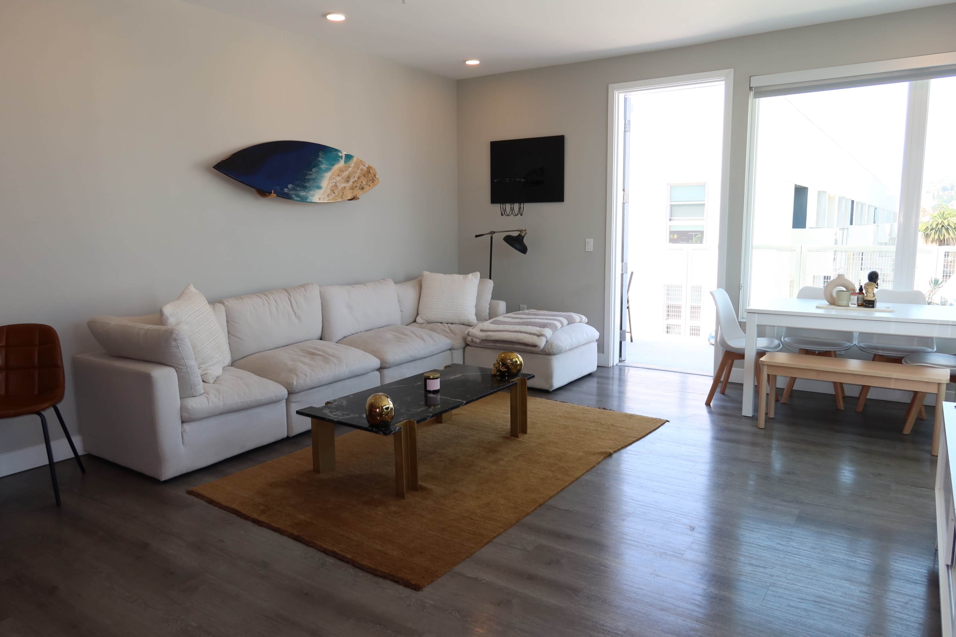 The image shows a modern living room with a white couch, a glass coffee table, a rug, and a dining area featuring a circular table and chairs next to a large window.