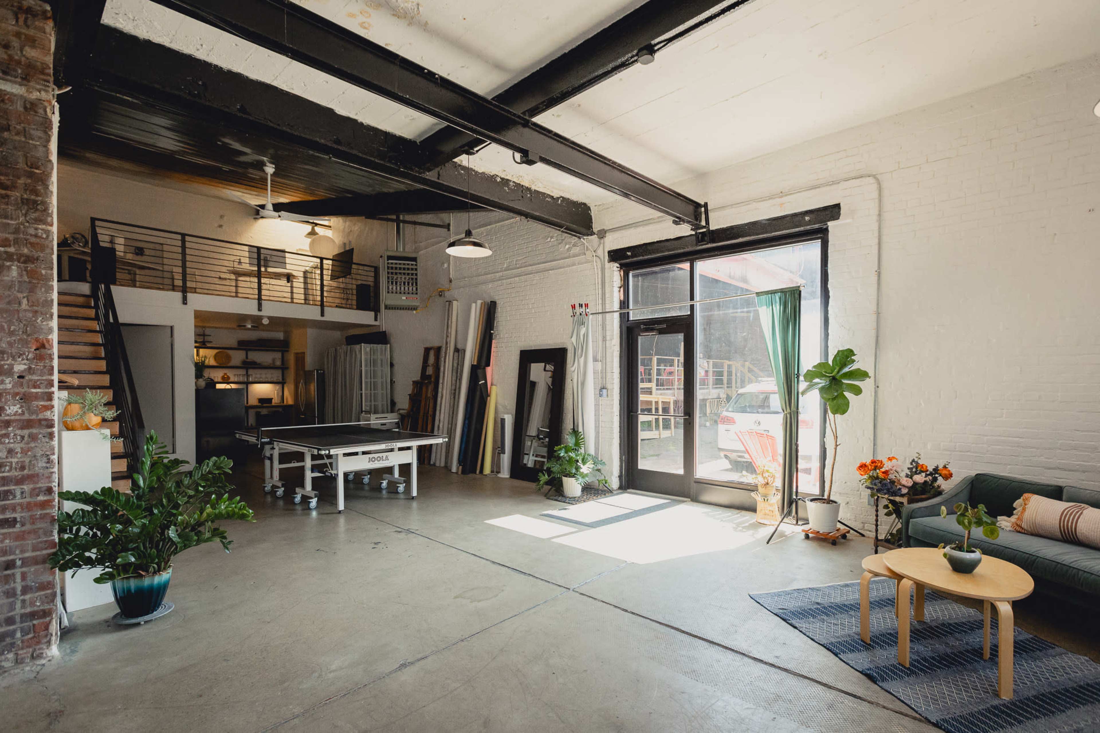 A spacious, industrial-style studio with a large open area, a kitchenette in the background, and natural light streaming through floor-to-ceiling windows.