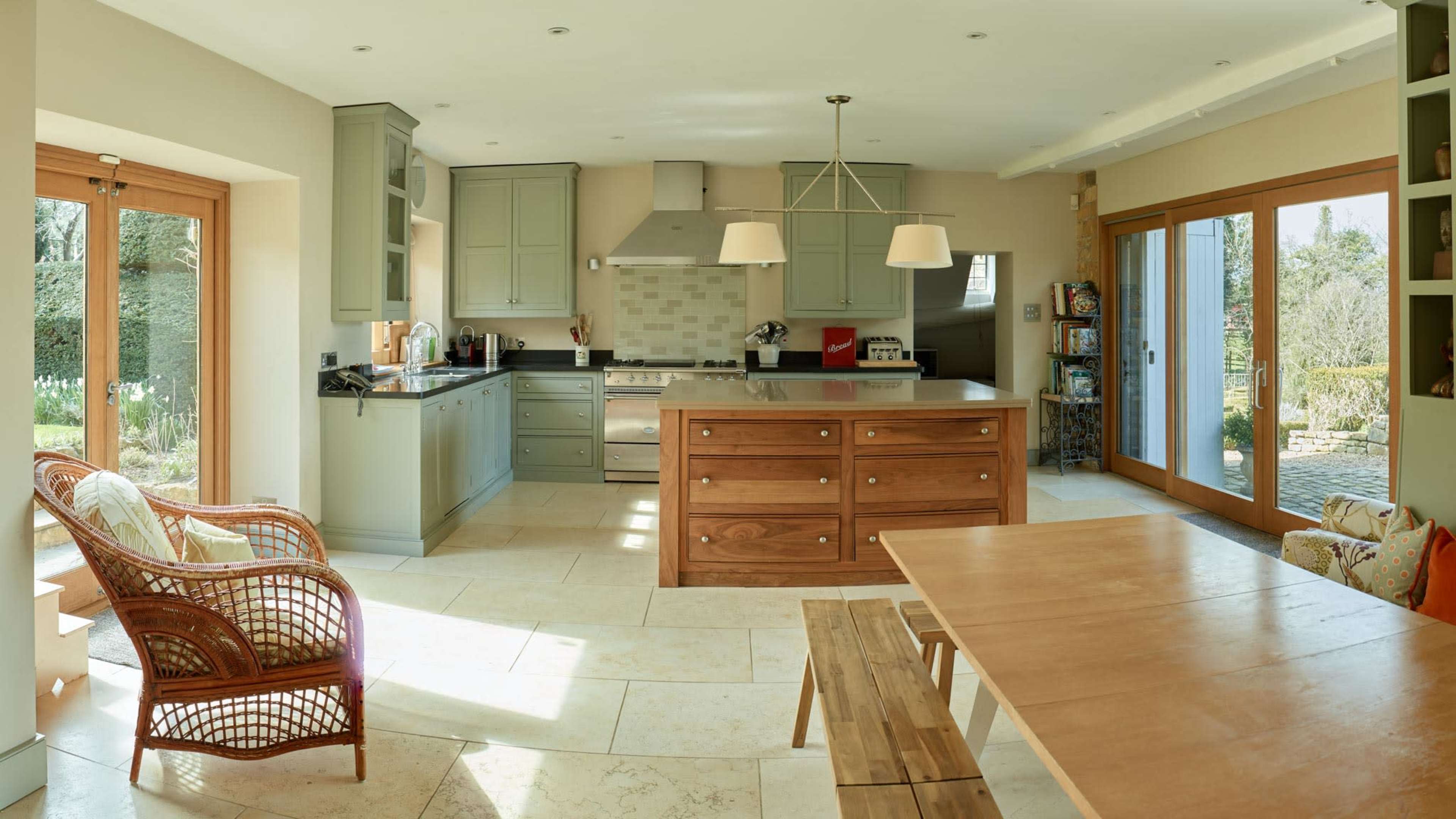 The image depicts a spacious kitchen with modern cabinetry, a large island, a dining table, and glass doors that open to a garden.