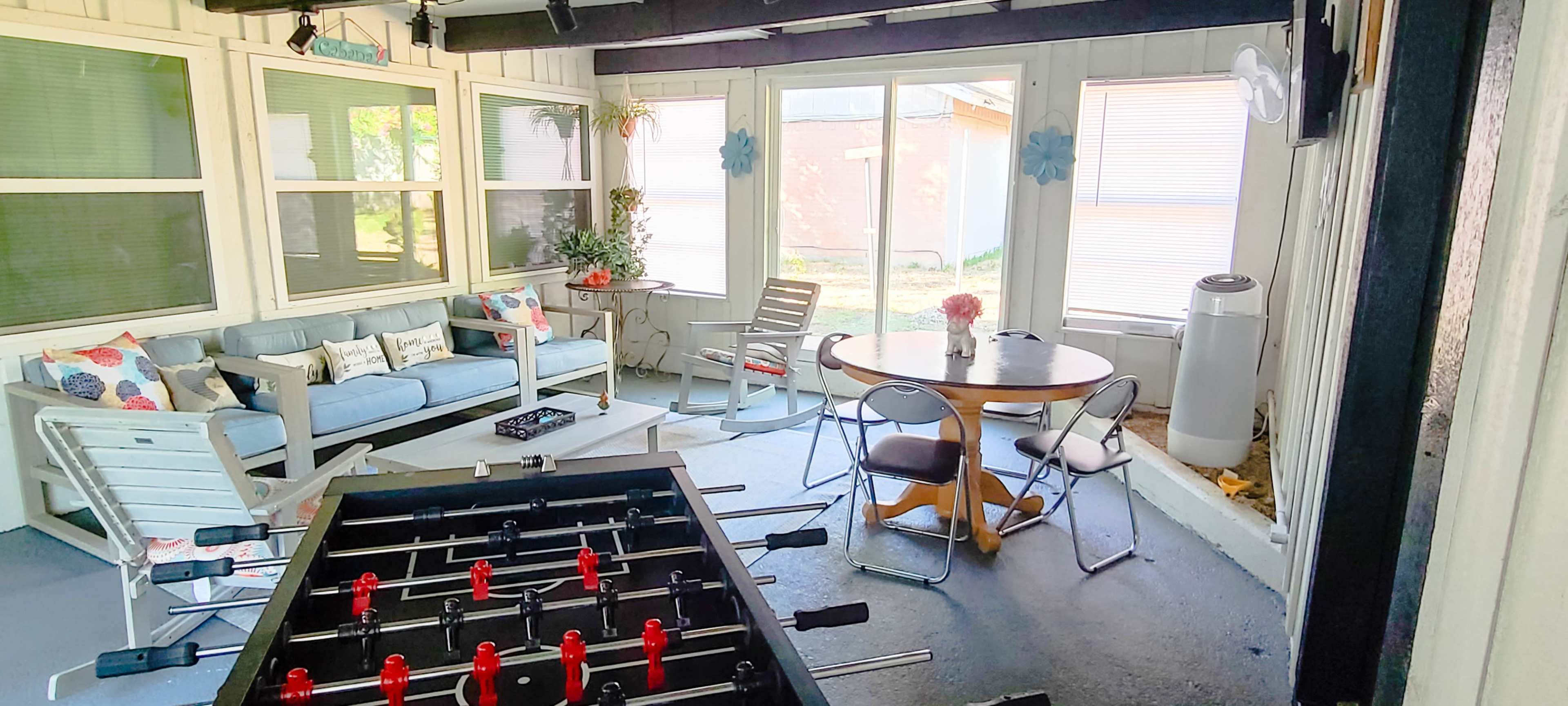 A screened-in porch with a seating area, a foosball table, and a dining table surrounded by chairs.