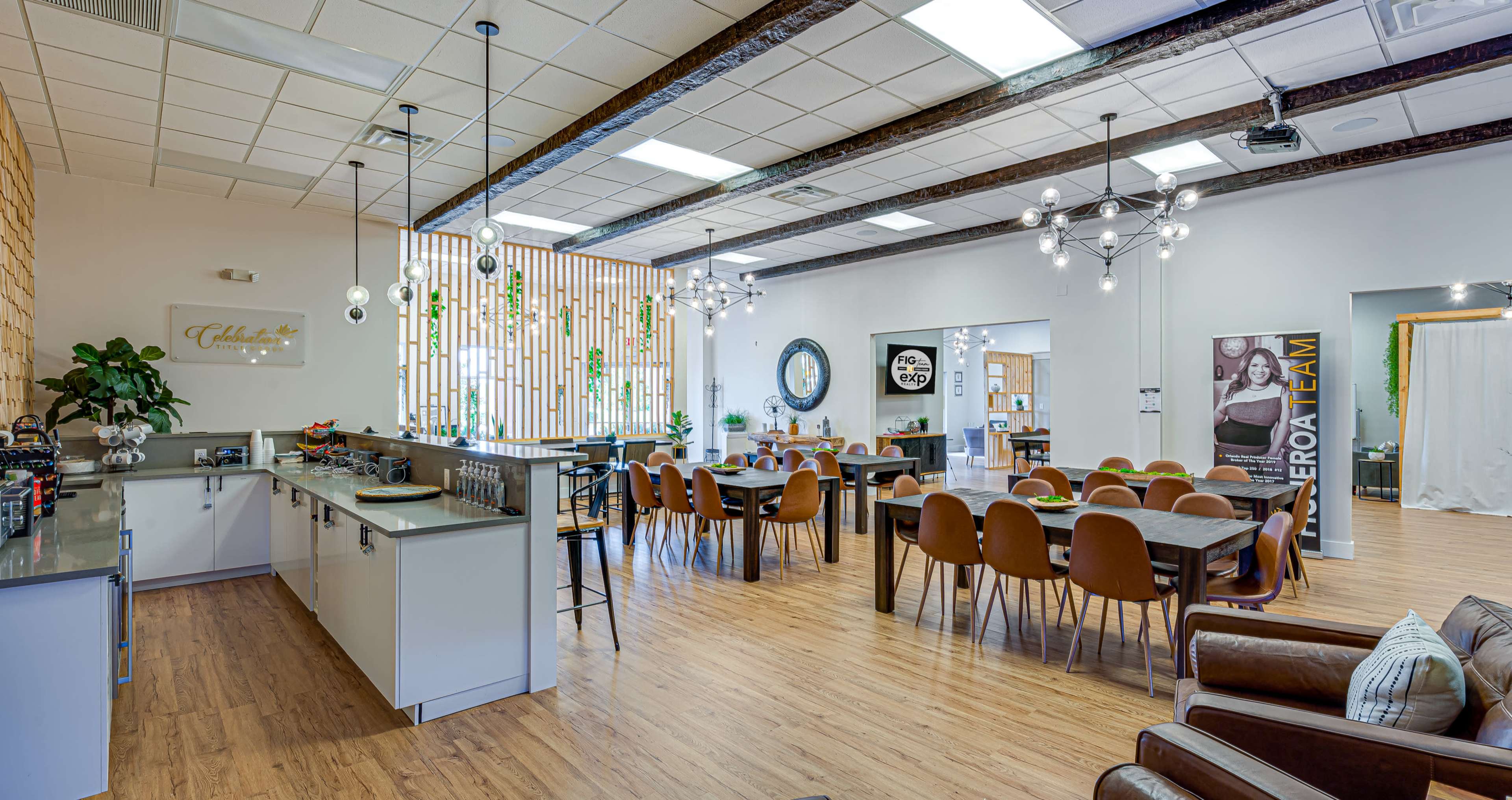 A spacious, well-lit interior of a modern office or communal space featuring a dining area with tables and chairs, a kitchen counter, and decorative plants.