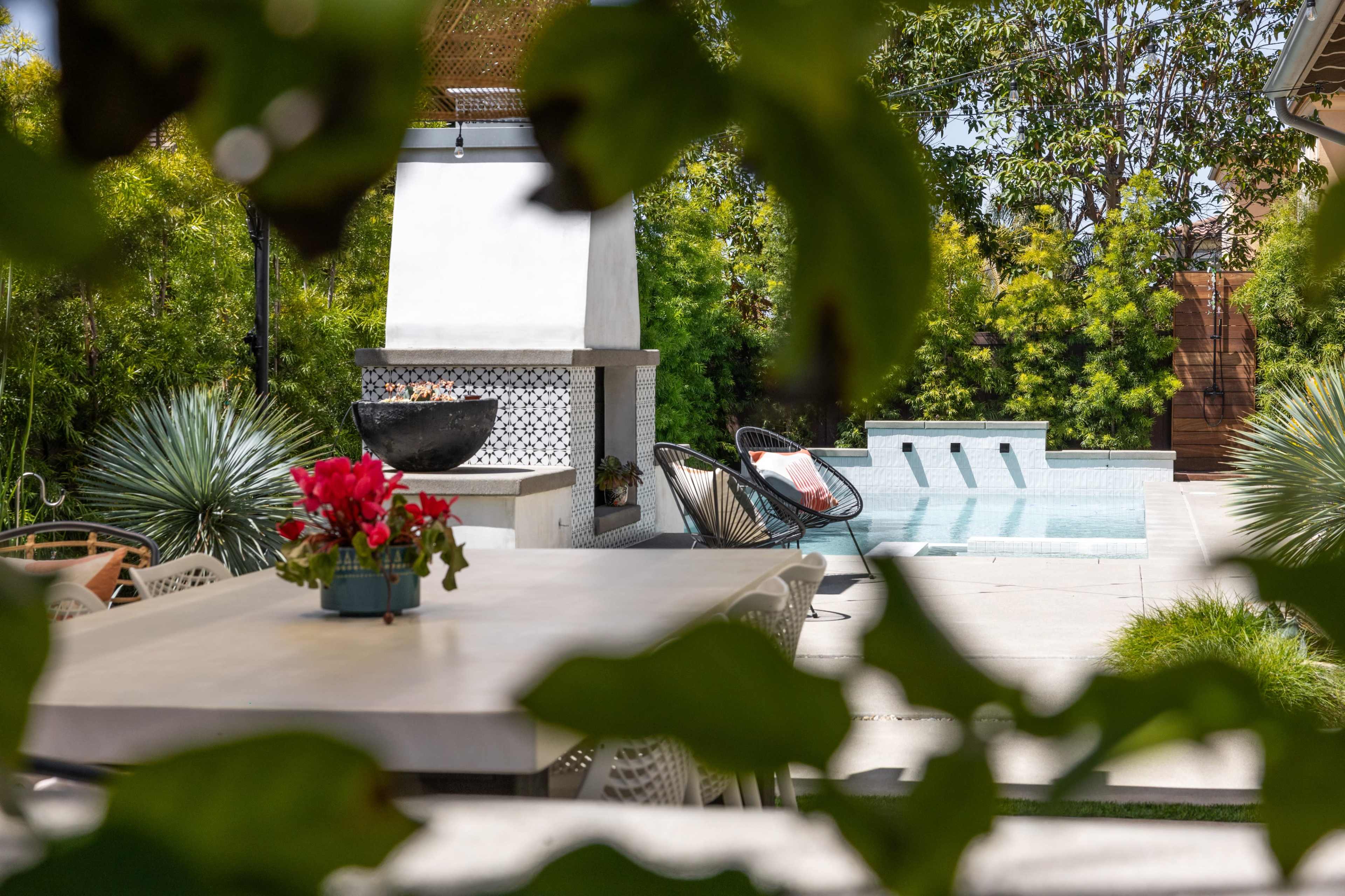 A patio features a dining table and a pool surrounded by greenery and a barbecue area.