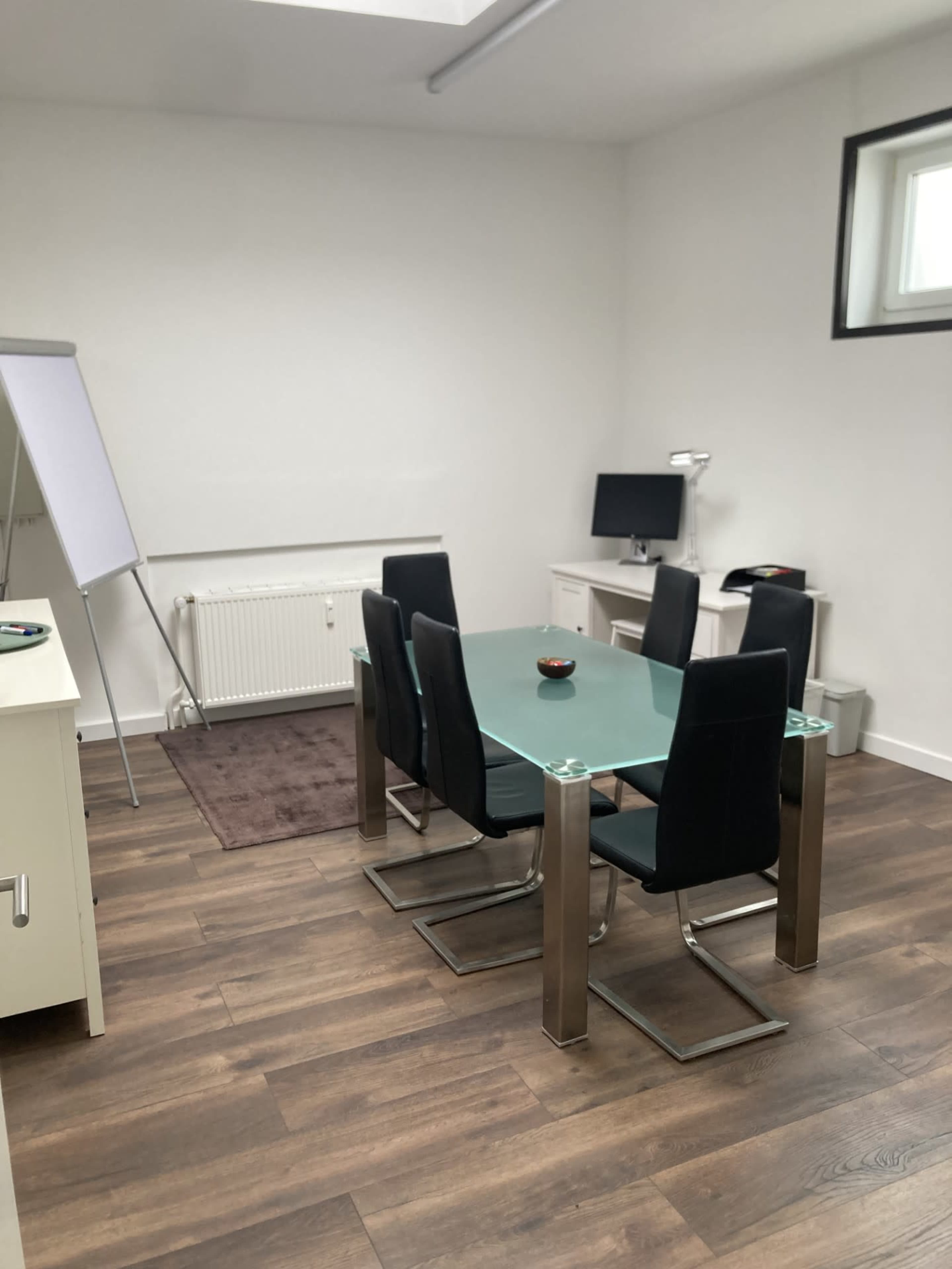 The image shows a modern office space with a glass table and four black chairs, a whiteboard, and a computer setup against a white wall.