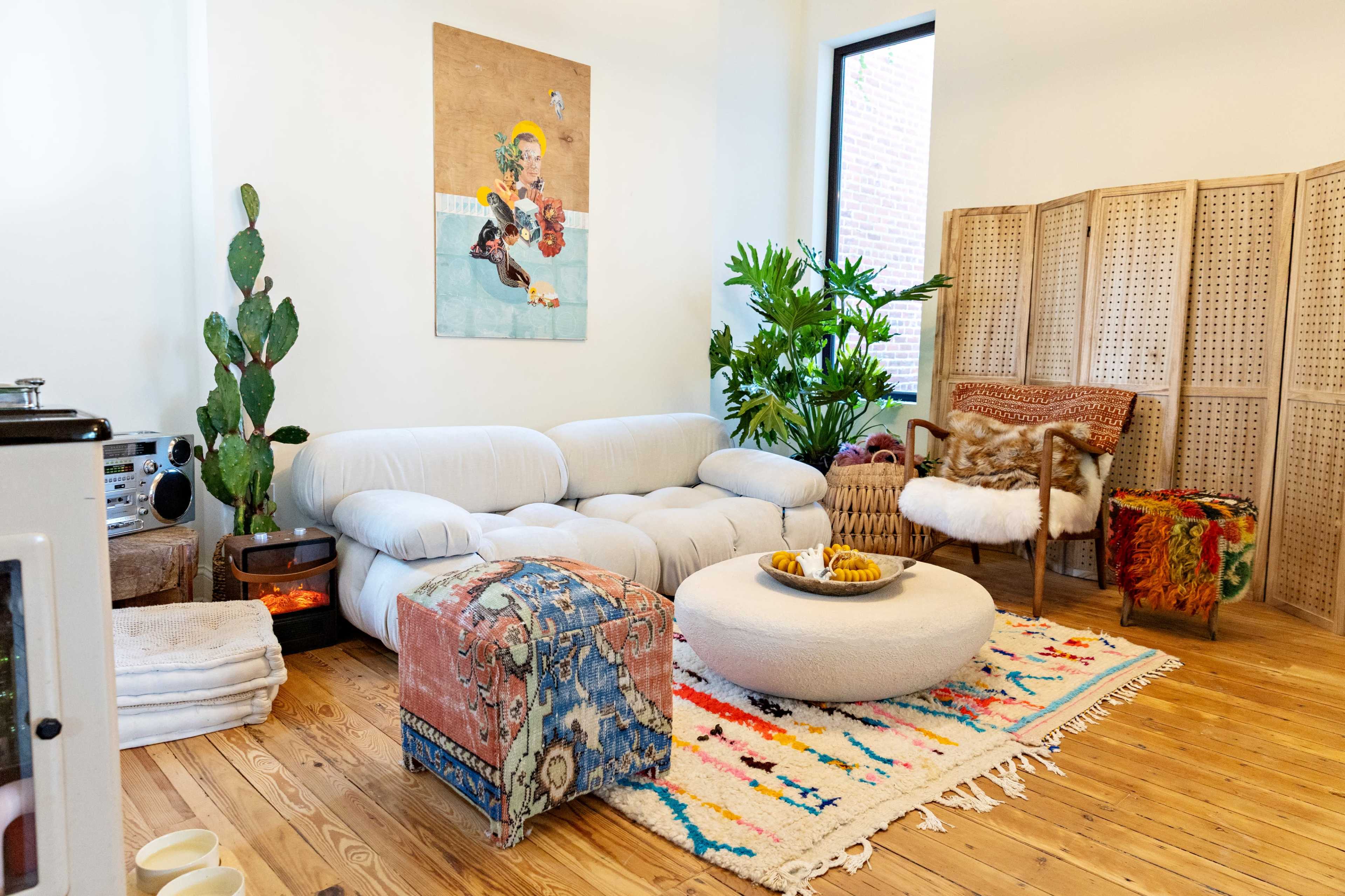 The scene features a cozy living room with a modern white sectional sofa, a circular coffee table, colorful rugs, indoor plants, and a decorative screen in the background.