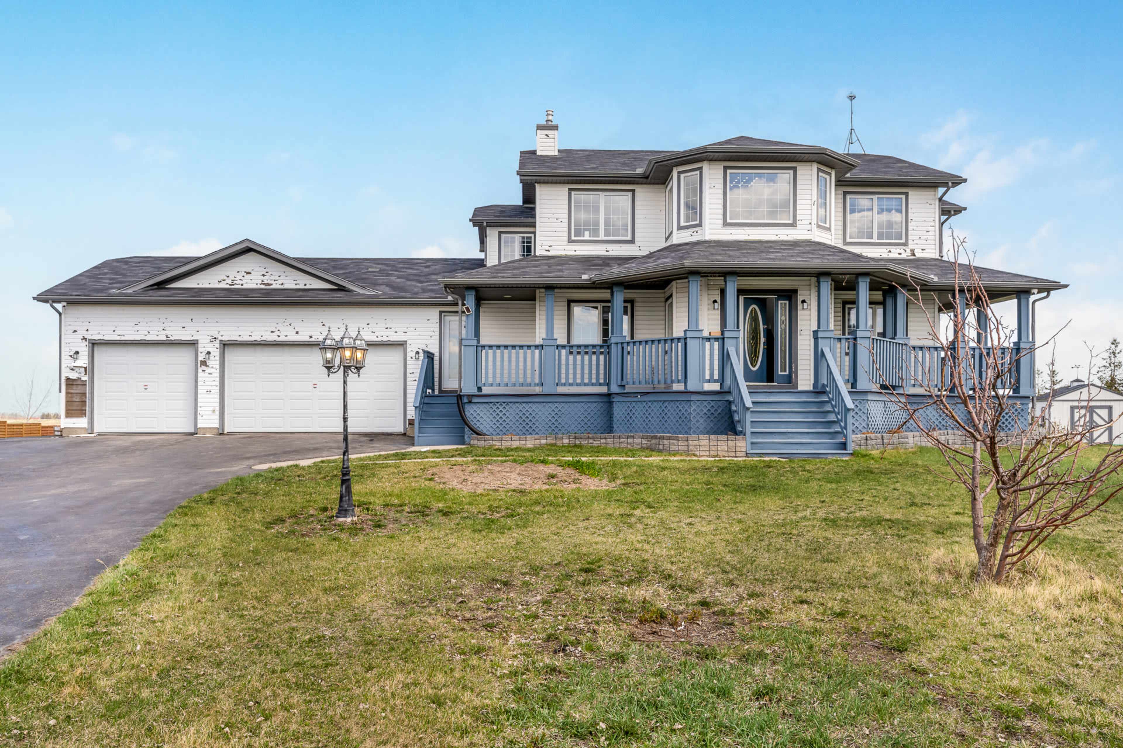 The image shows a large, two-story house with a wraparound porch, a circular driveway, and a detached garage on a grassy lot.