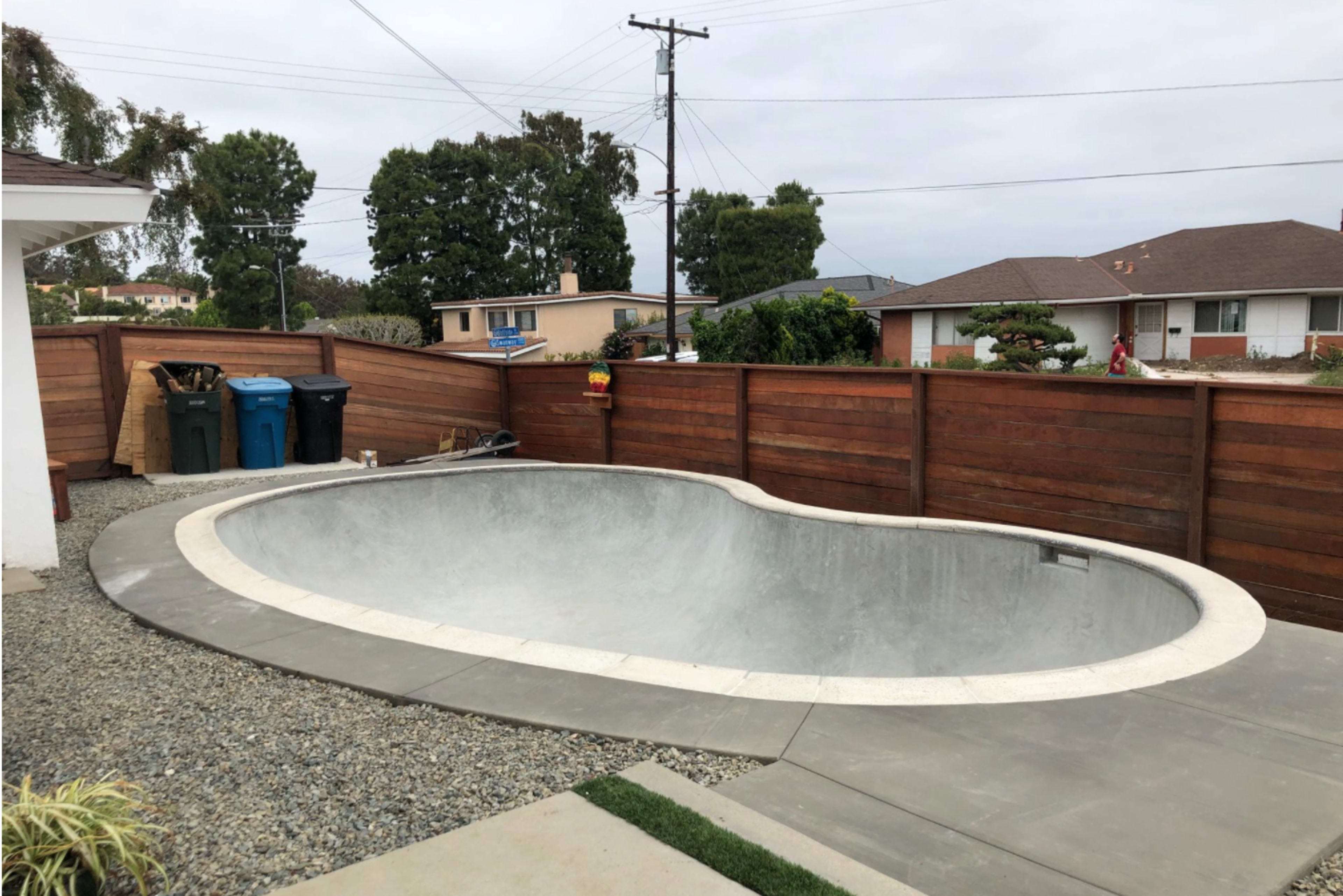 An empty, unfinished concrete swimming pool surrounded by a fenced yard with a gravel pathway and garbage bins nearby.