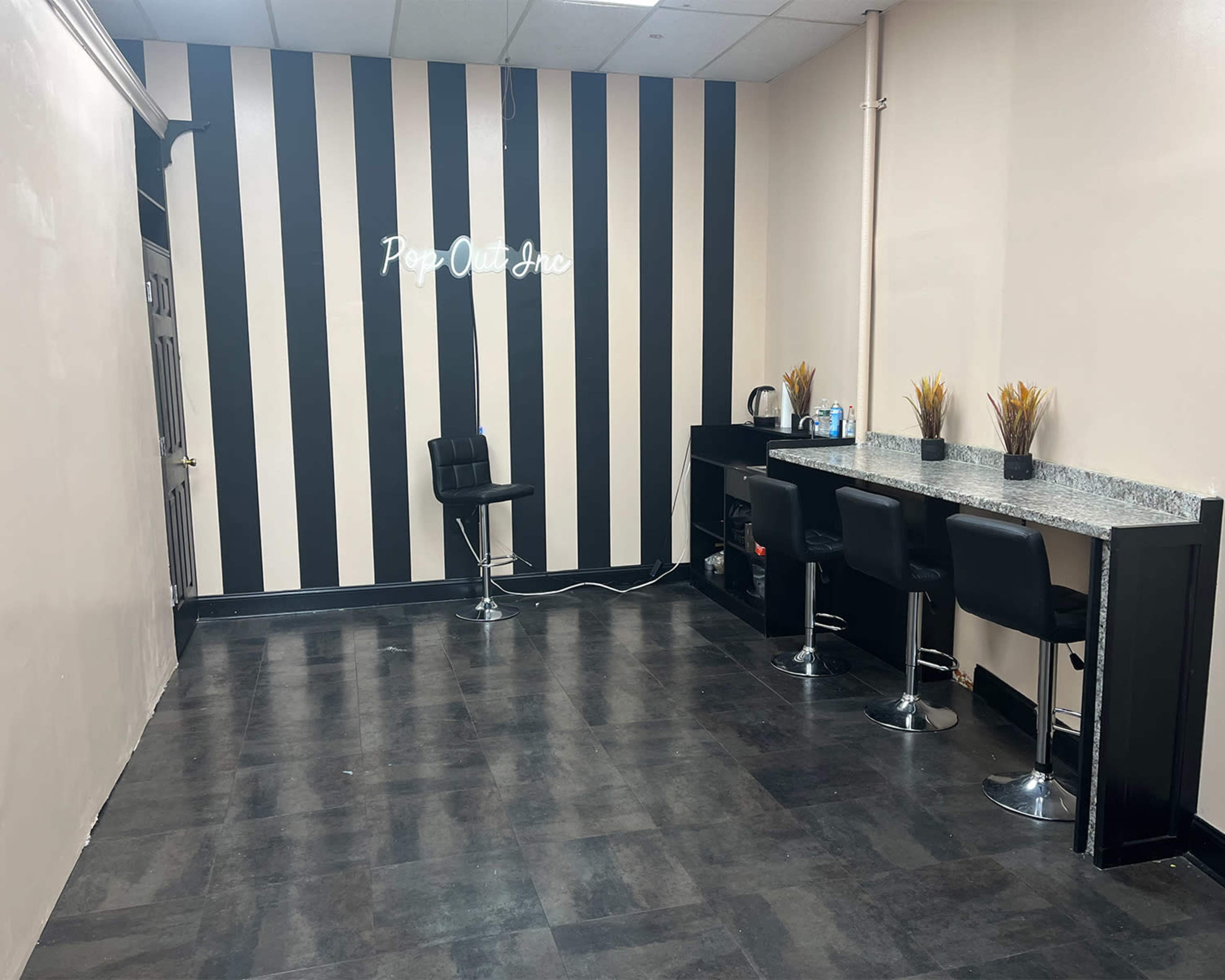 A small room with black and white striped walls, featuring a countertop with four black barstools and a neon sign that reads "Pop Out."