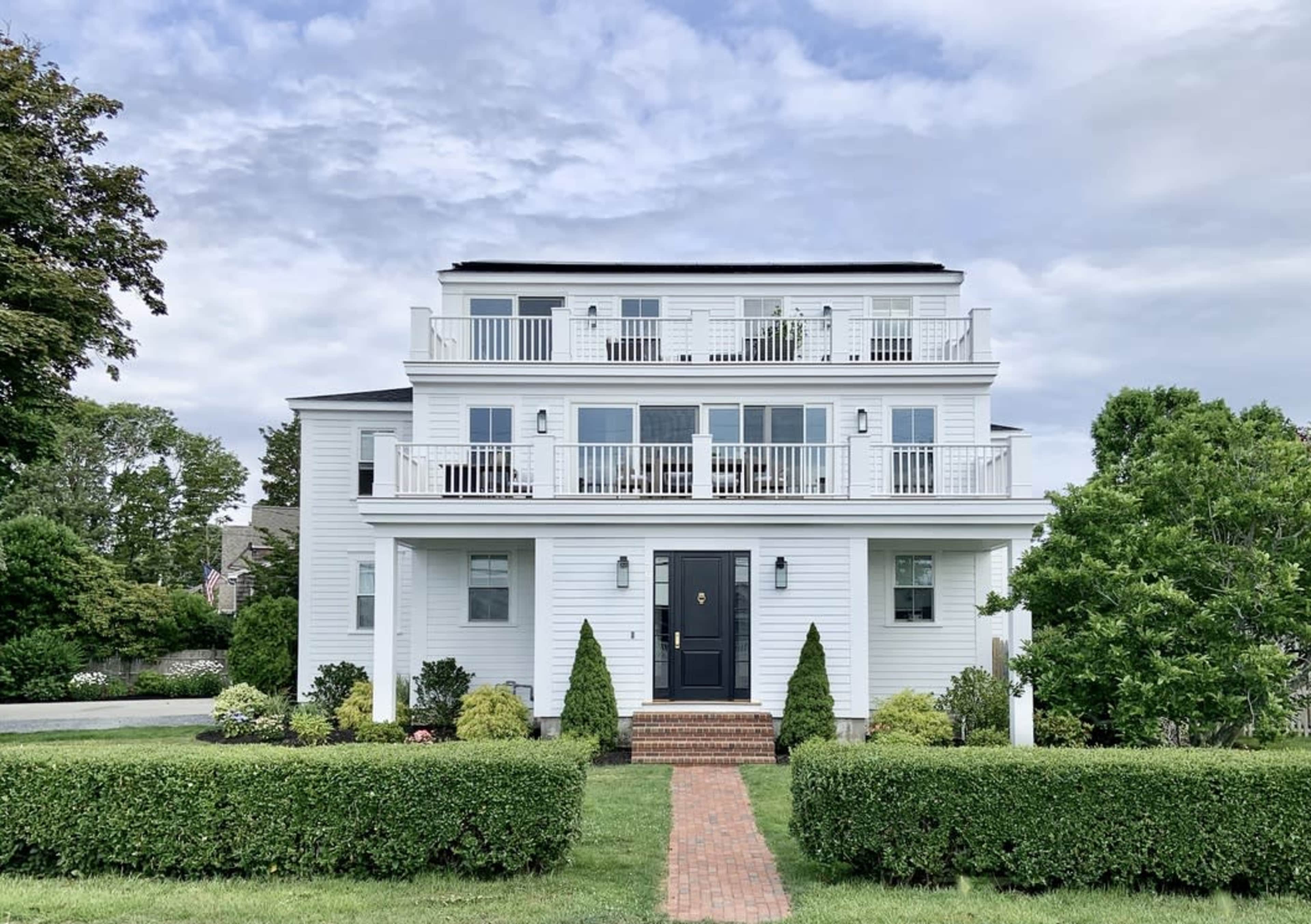 A three-story white house with balconies on each floor is flanked by neatly trimmed hedges and a brick pathway leading to the front door.