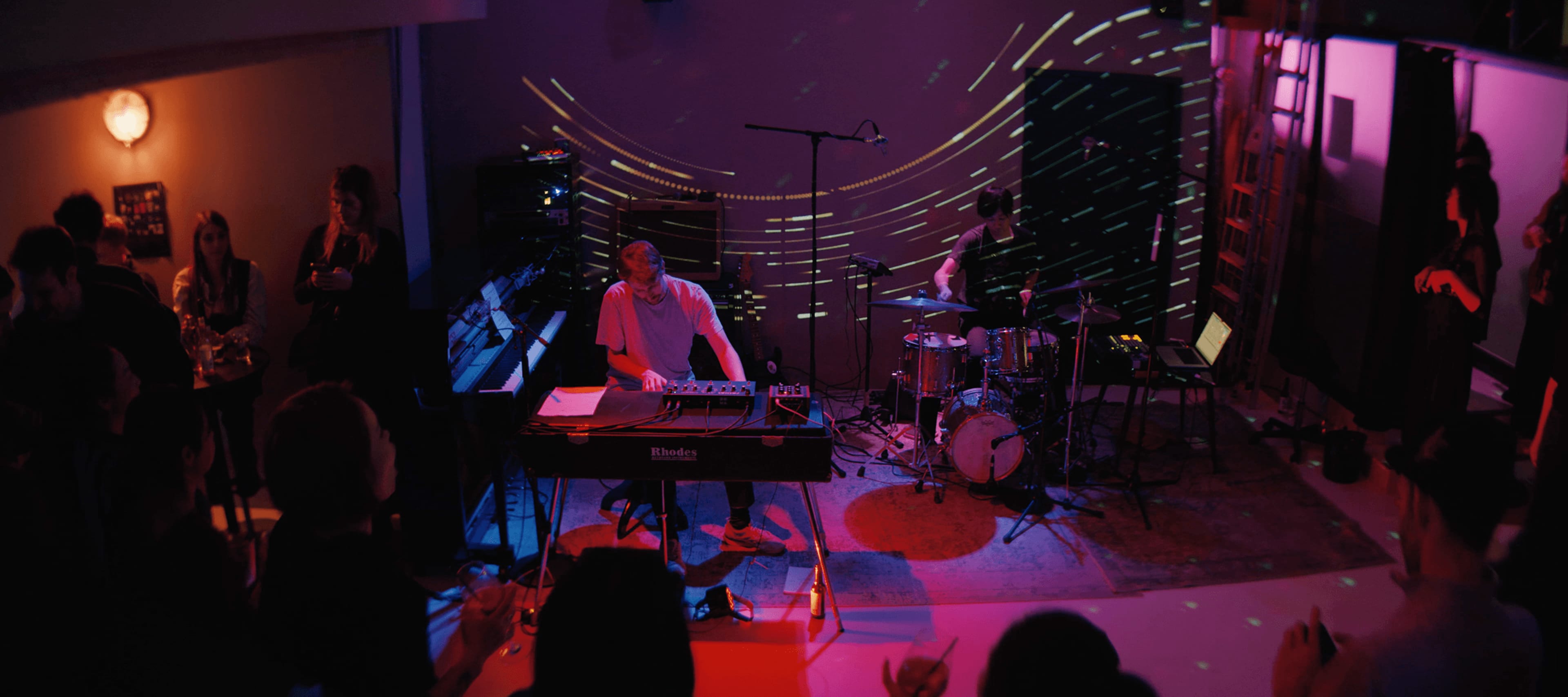 A live music performance is taking place in an ambient venue with colorful lights and a small audience facing the musicians at a keyboard and drums.
