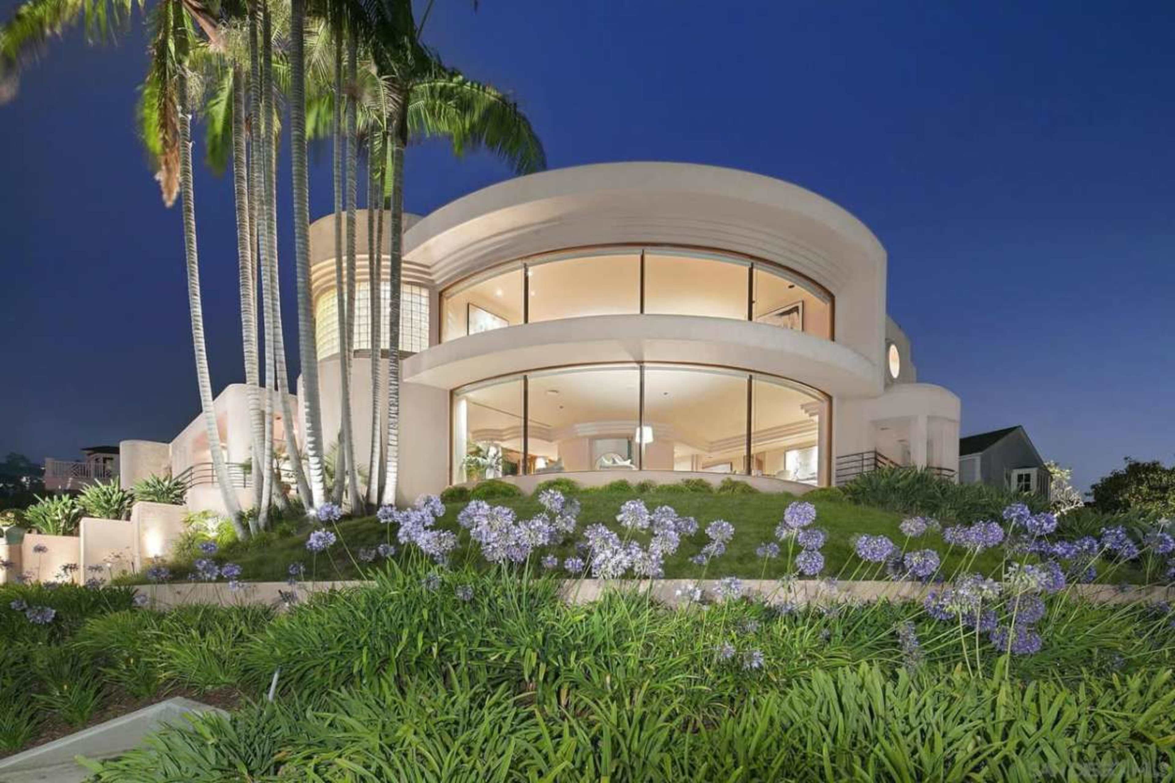 A modern, round-shaped house with large windows is set among lush greenery and palm trees at dusk.