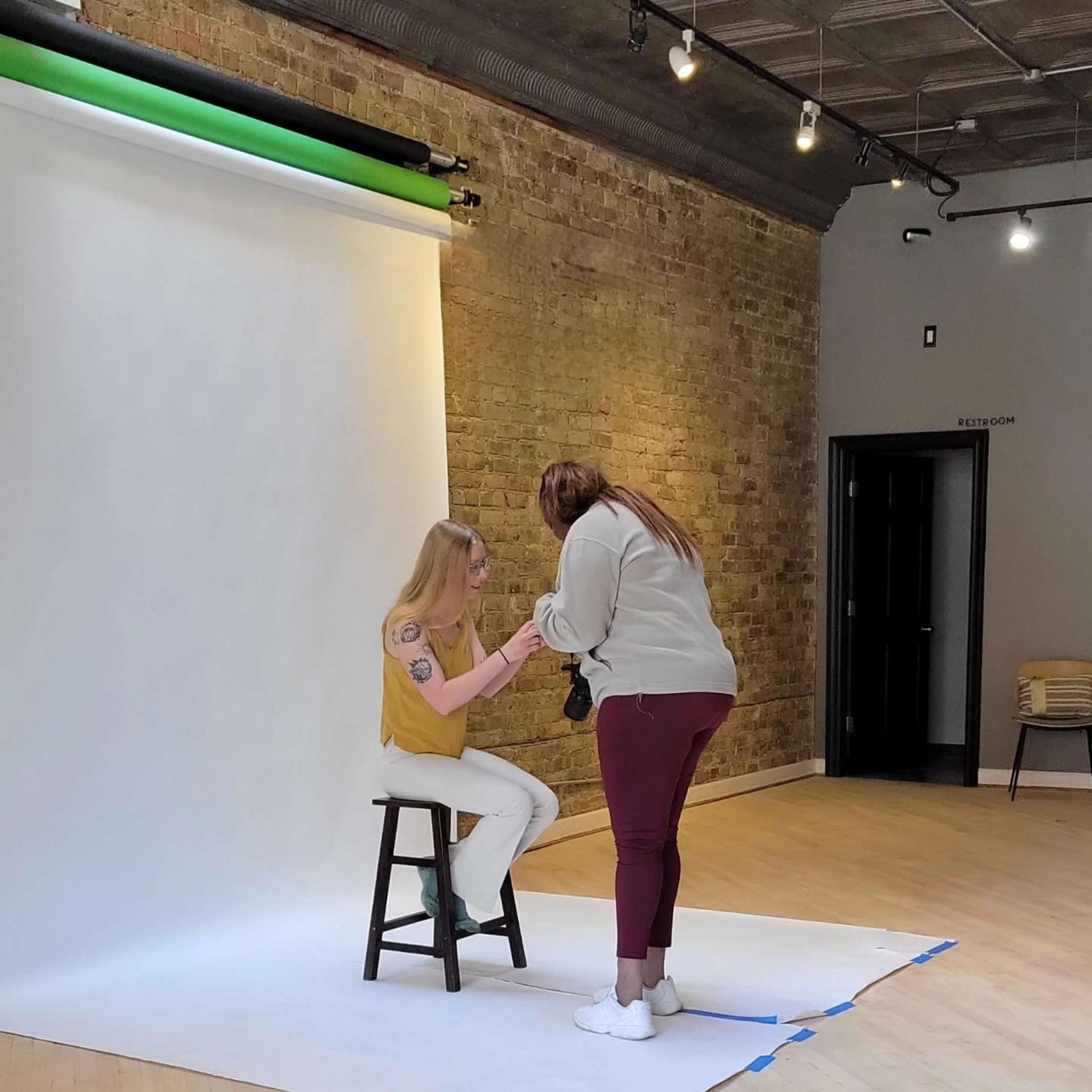 A woman sits on a stool in a photo studio while another woman adjusts her outfit.